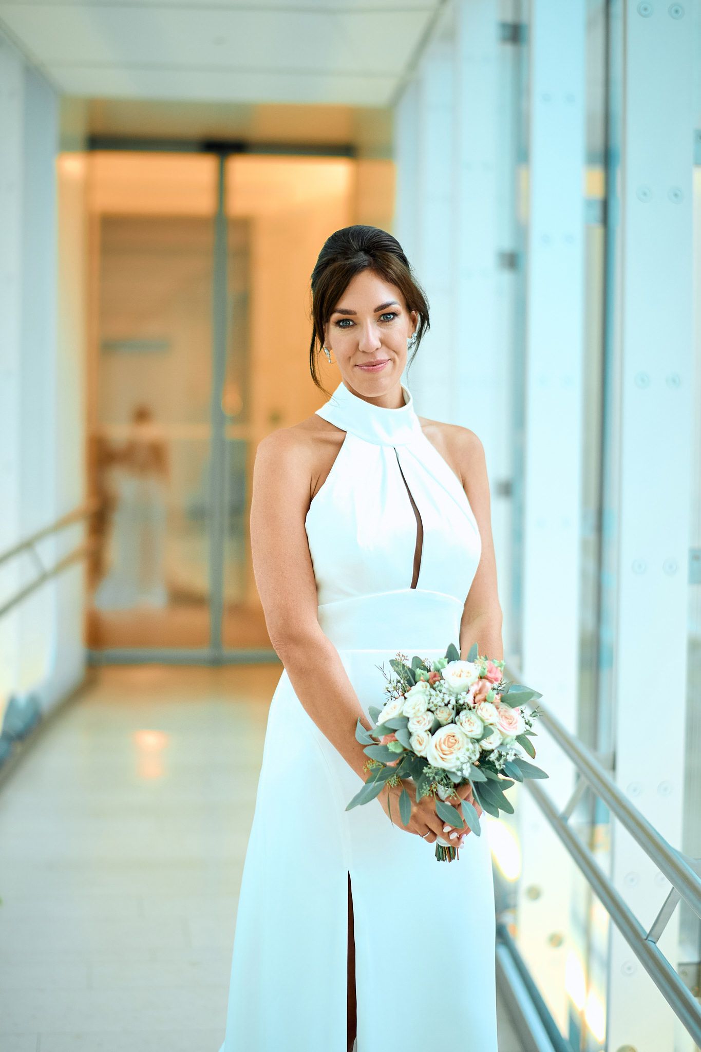 Elegant bride in a modern white dress poses in a sunlit corridor with a floral bouquet.