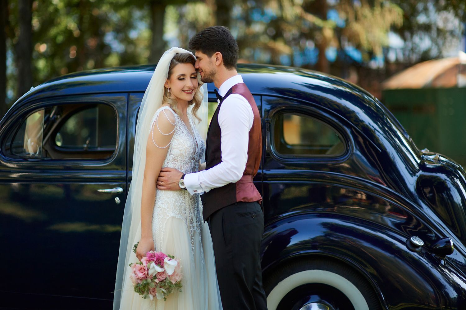 Newlyweds pose romantically by vintage black car during wedding photoshoot.