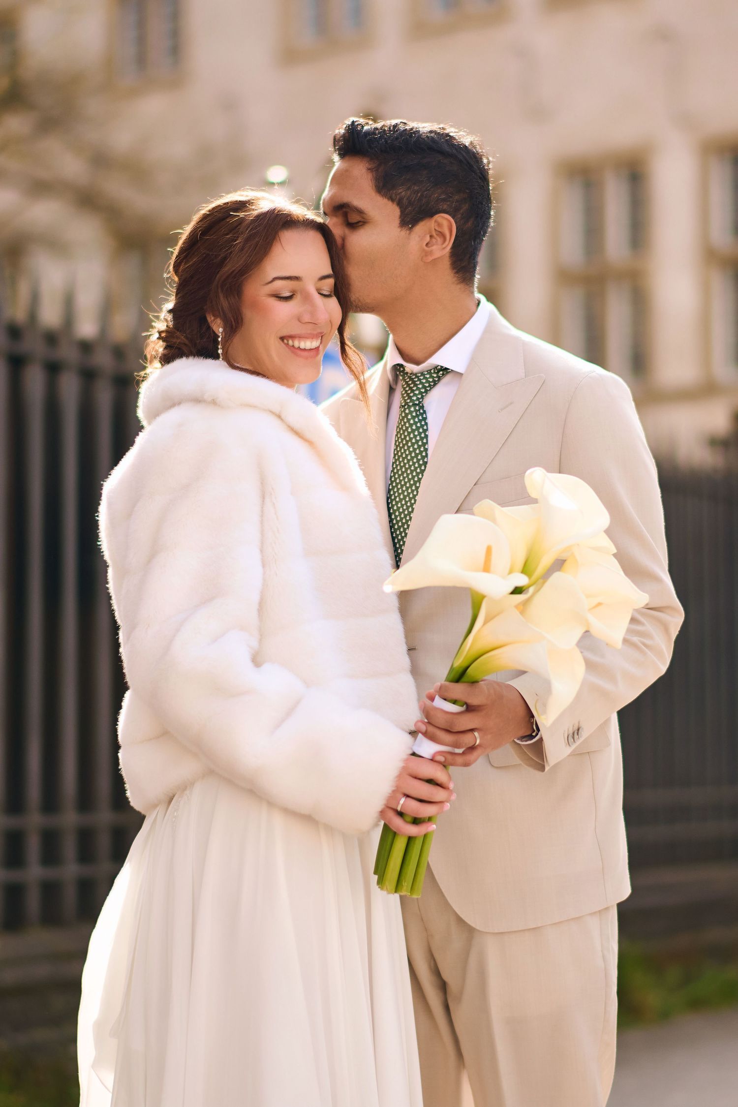Groom in beige suit kisses bride wearing white fur coat holding calla lily bouquet outdoors on wedding day.
