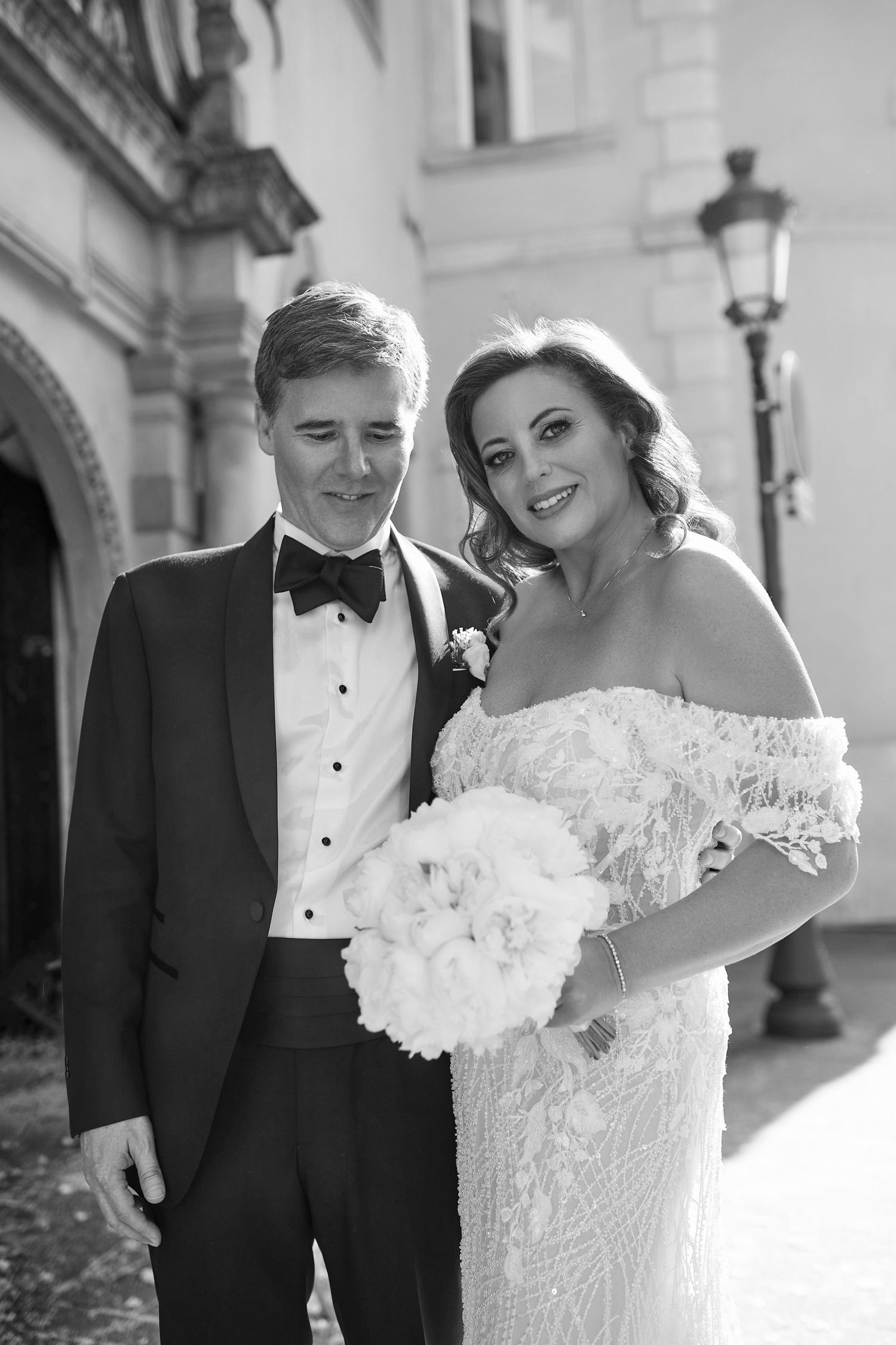 Black and white wedding portrait of smiling bride in off-shoulder gown holding bouquet with groom in tuxedo.