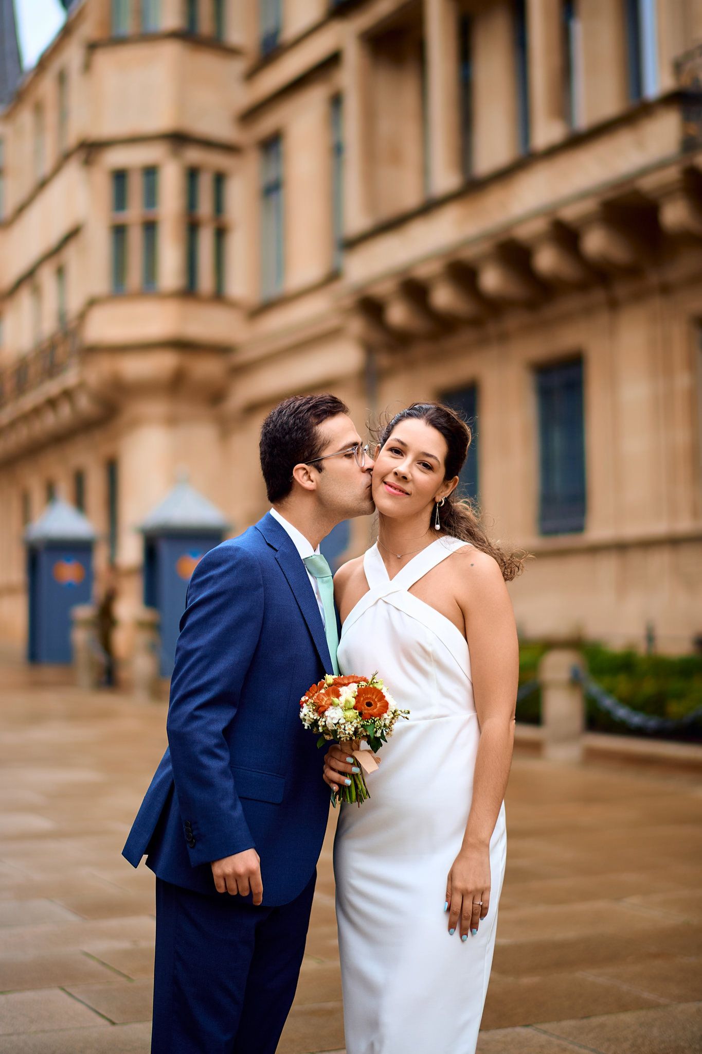 Couple in formal attire shares intimate moment against stone building backdrop.