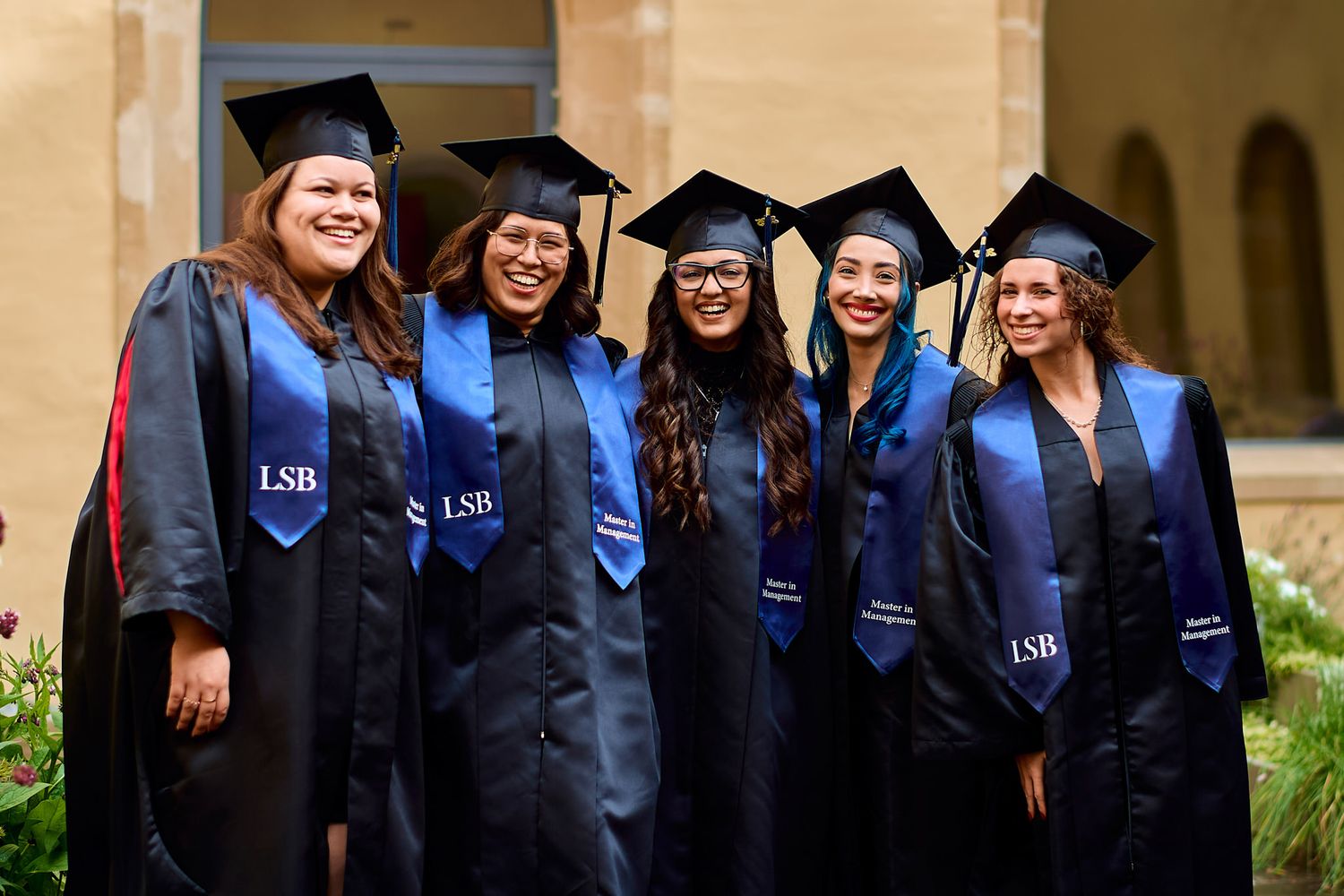 Students wearing graduation regalia smile and celebrate on campus.