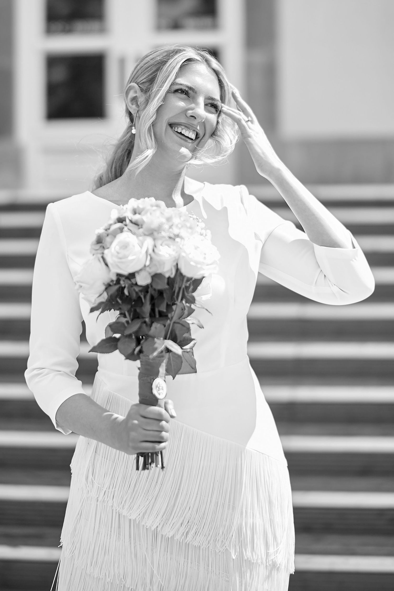 A joyful bride in a white fringe dress holds a bouquet of roses while standing on outdoor steps.