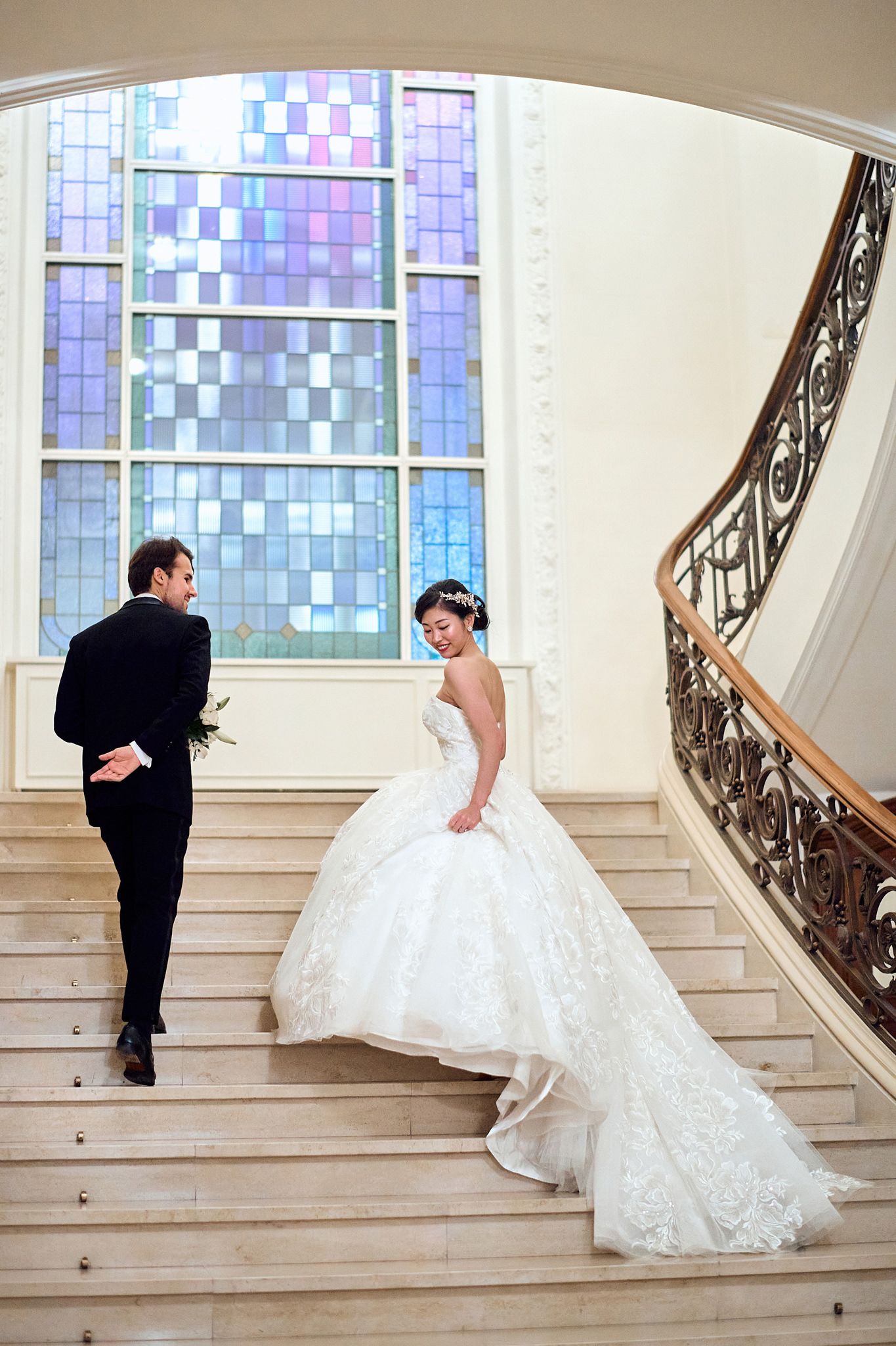 A couple shares their first look moment on an elegant staircase with stained glass windows.