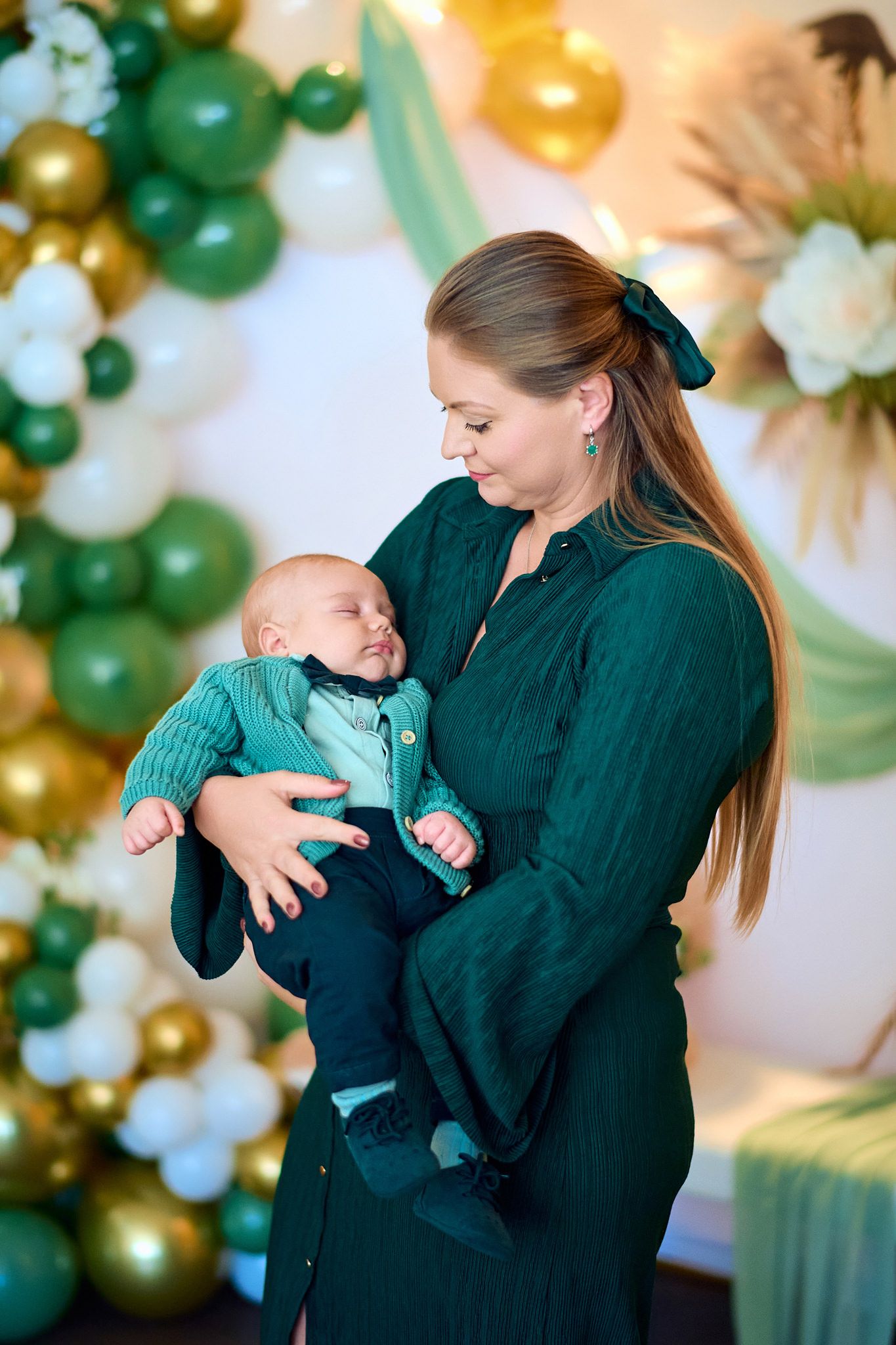 Mother in green sweater holds and comforts baby against balloon backdrop.