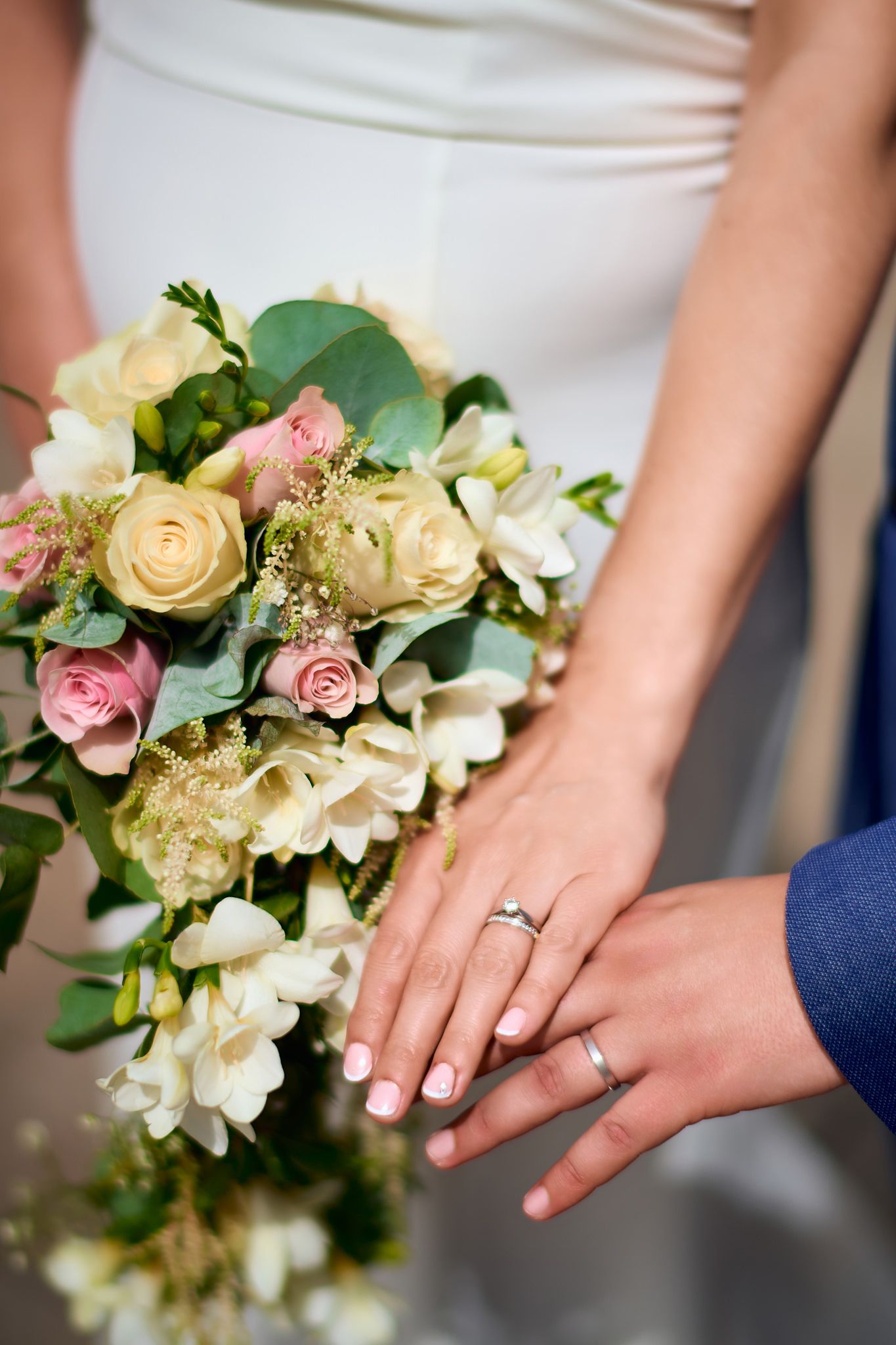 Close-up of wedding rings and hands against a beautiful floral bouquet with roses and greenery.