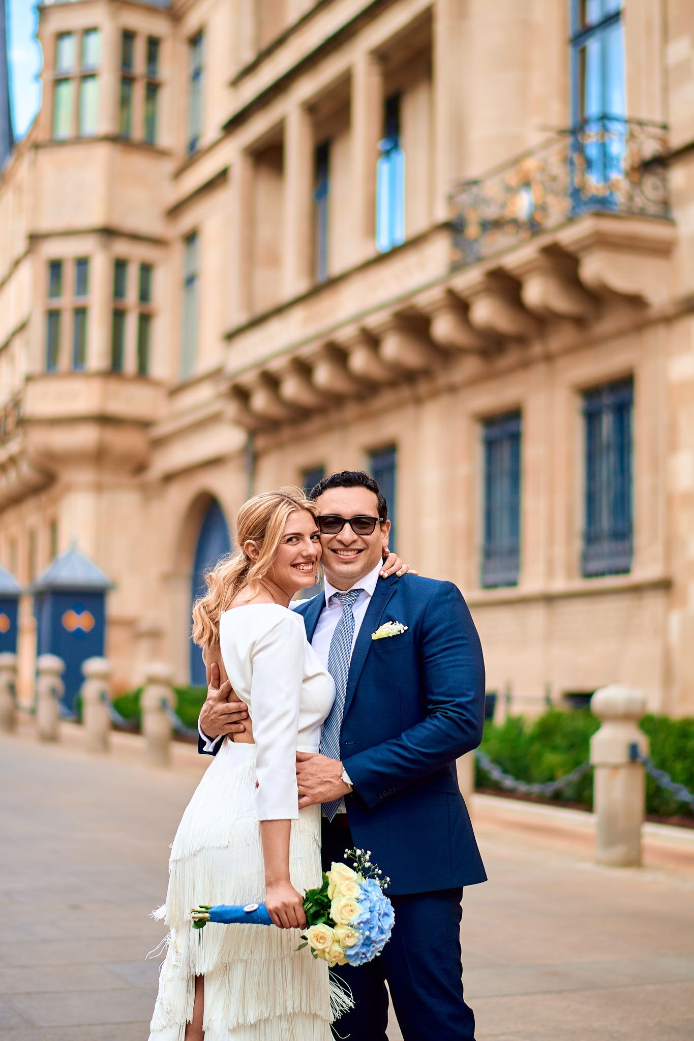 A couple embraces outdoors near a classical stone building on a sunny day.