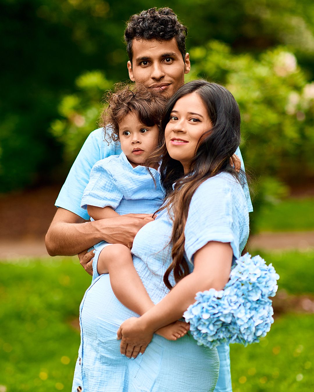 A family of three in matching blue outfits poses outdoors, with a pregnant woman holding blue hydrangeas in a lush green park.