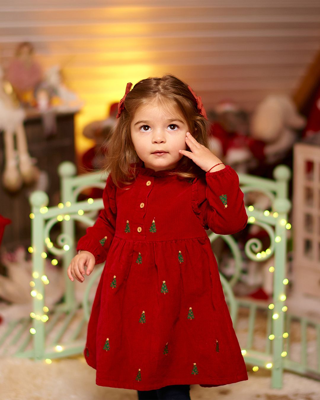 Toddler girl in red Christmas tree embroidered dress poses in festive holiday photo setting with warm lights.