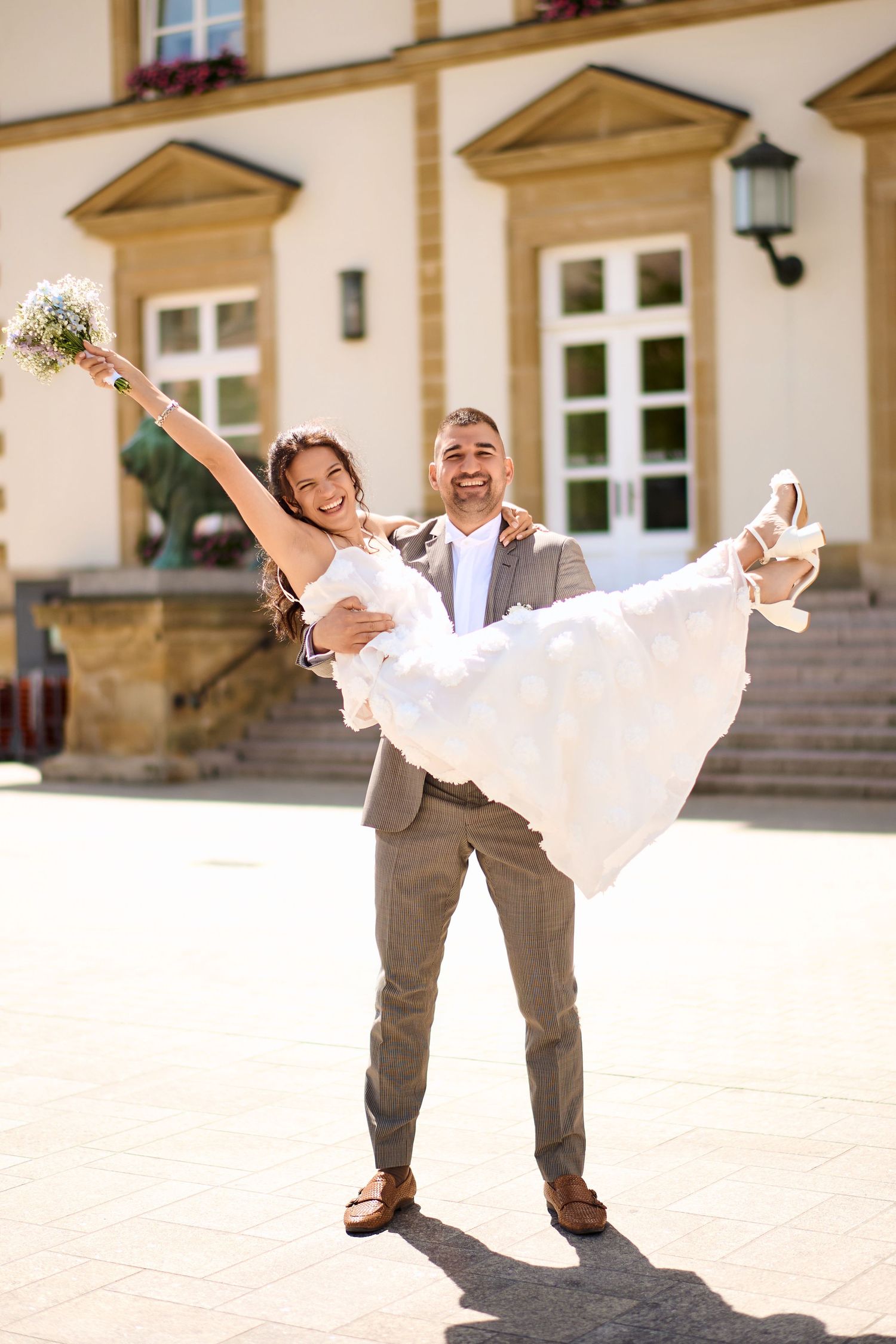 Groom in gray suit carries smiling bride in white dress outside sunny wedding venue.