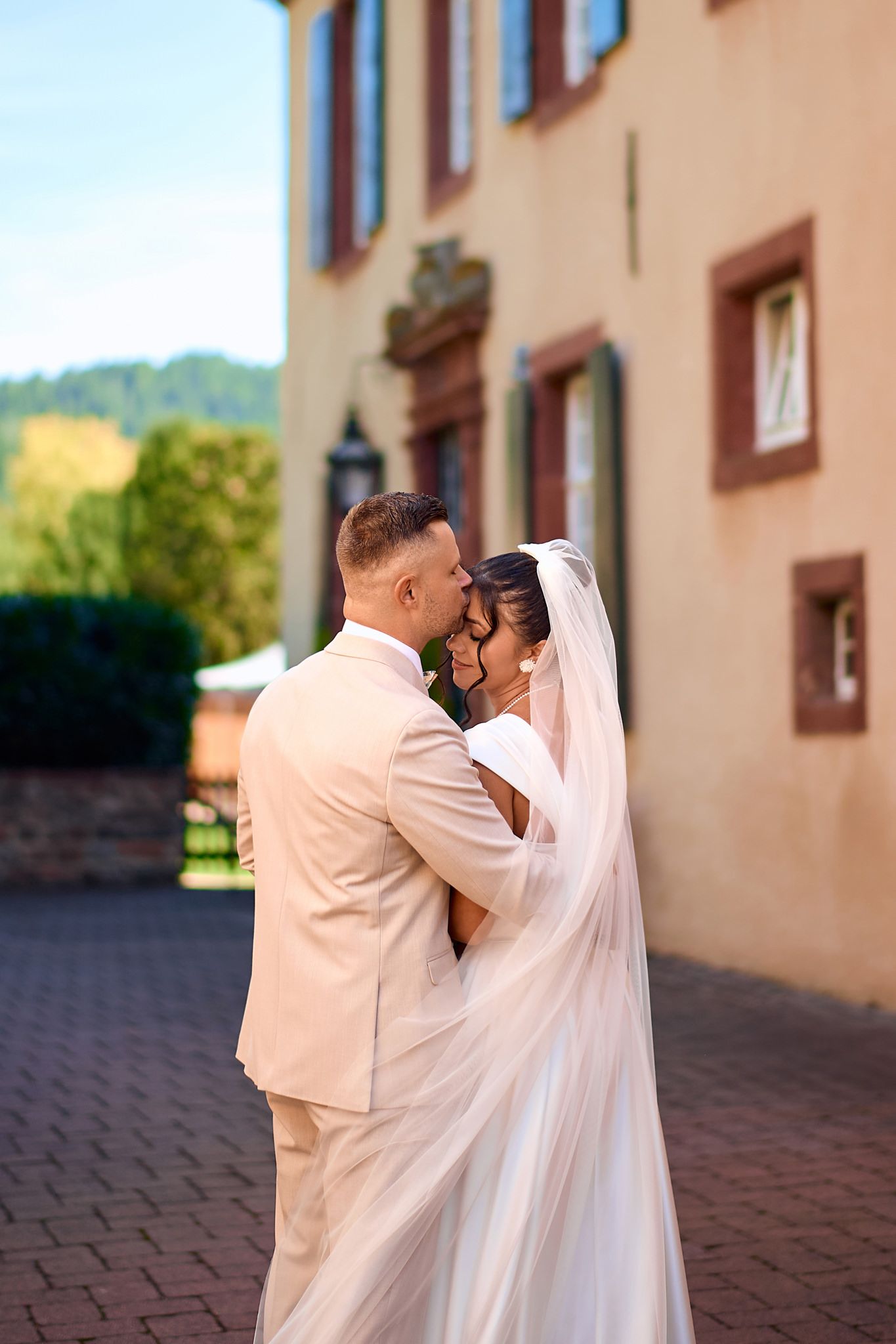 A couple in white wedding attire shares a romantic moment on cobblestone pavement.