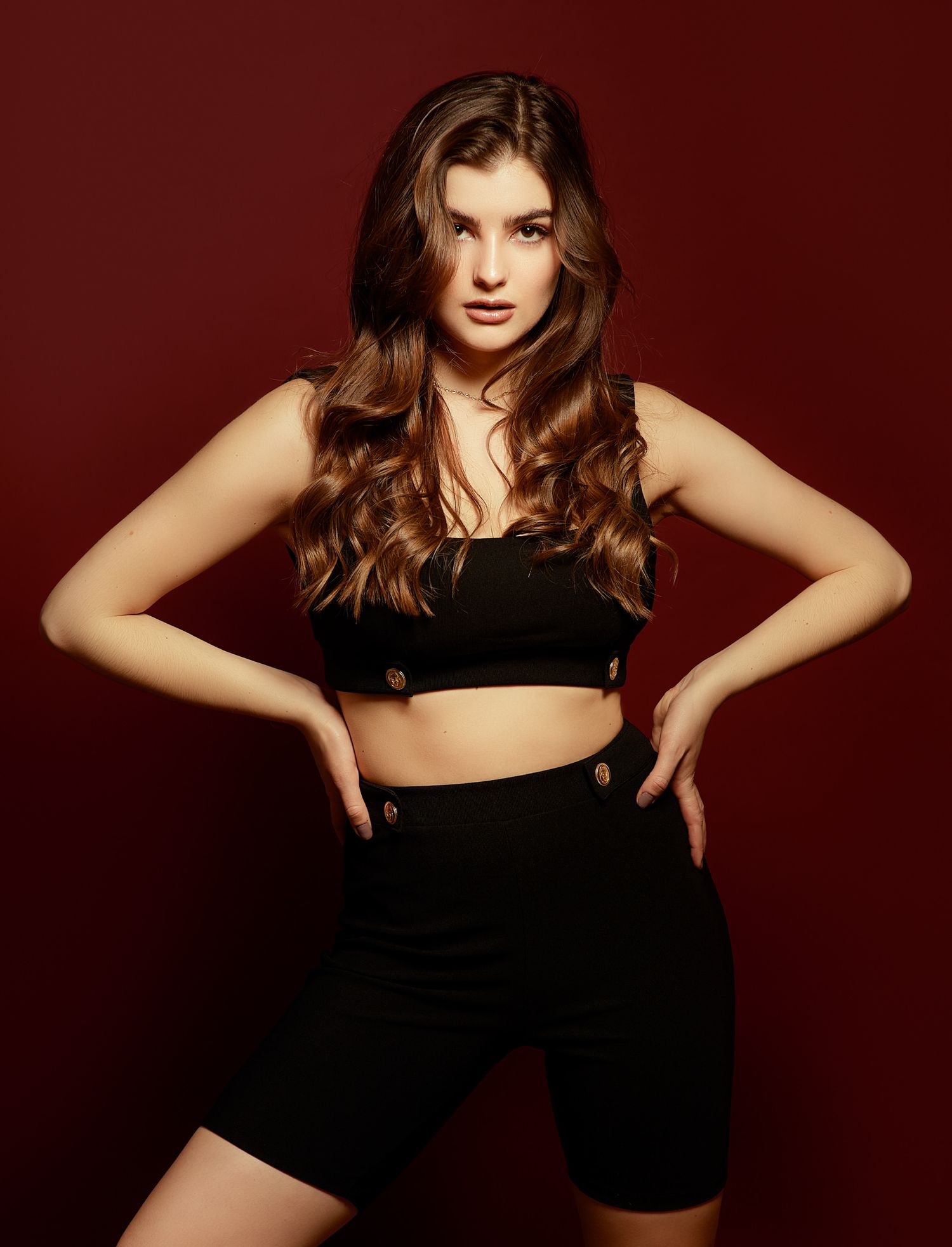 Athletic pose in black crop top and shorts against dark red backdrop.