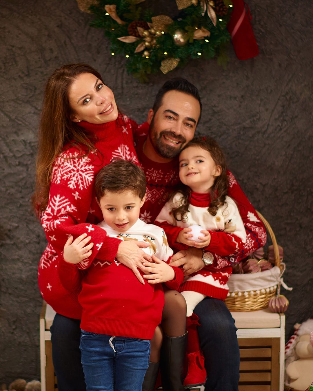Family of four in matching red Christmas sweaters posing for a holiday portrait with festive wreath backdrop.