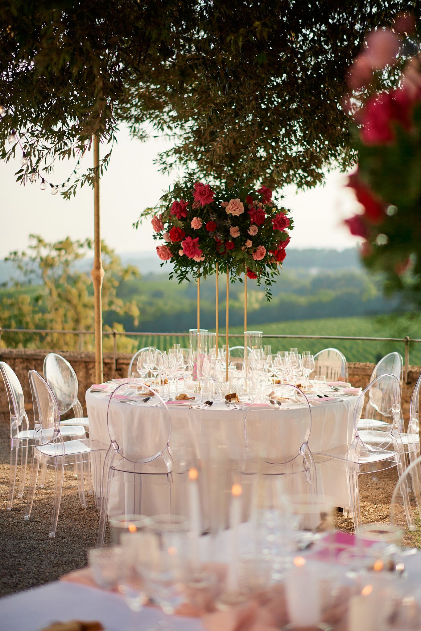 Romantic outdoor reception setup with white linens and red floral centerpieces against rolling hills.