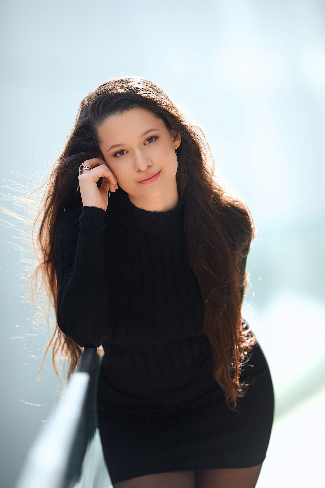 Woman with long dark hair wearing black turtleneck in bright studio setting.