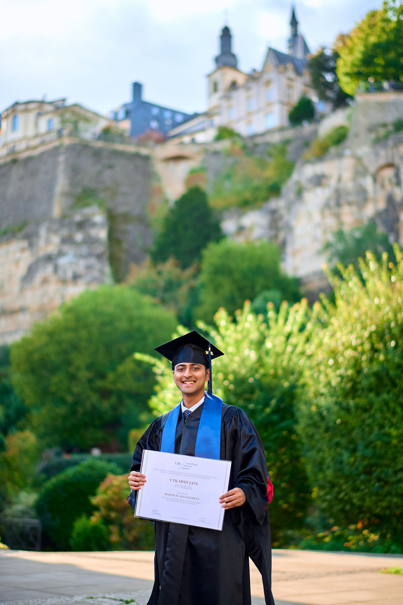 Graduate in academic regalia poses with diploma against scenic castle backdrop and greenery.