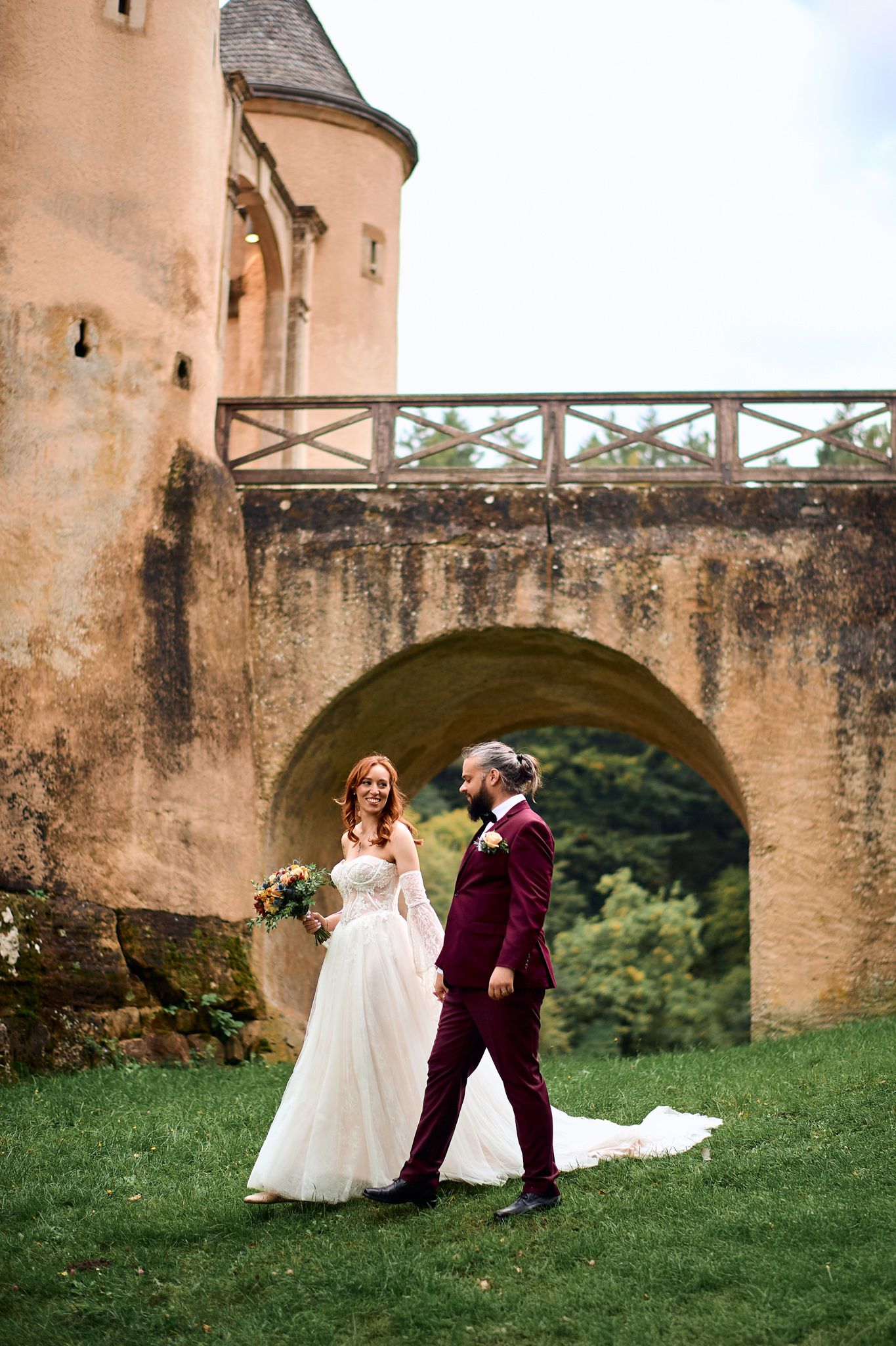 Wedding couple poses playfully on grass near historic stone castle wall and archway.