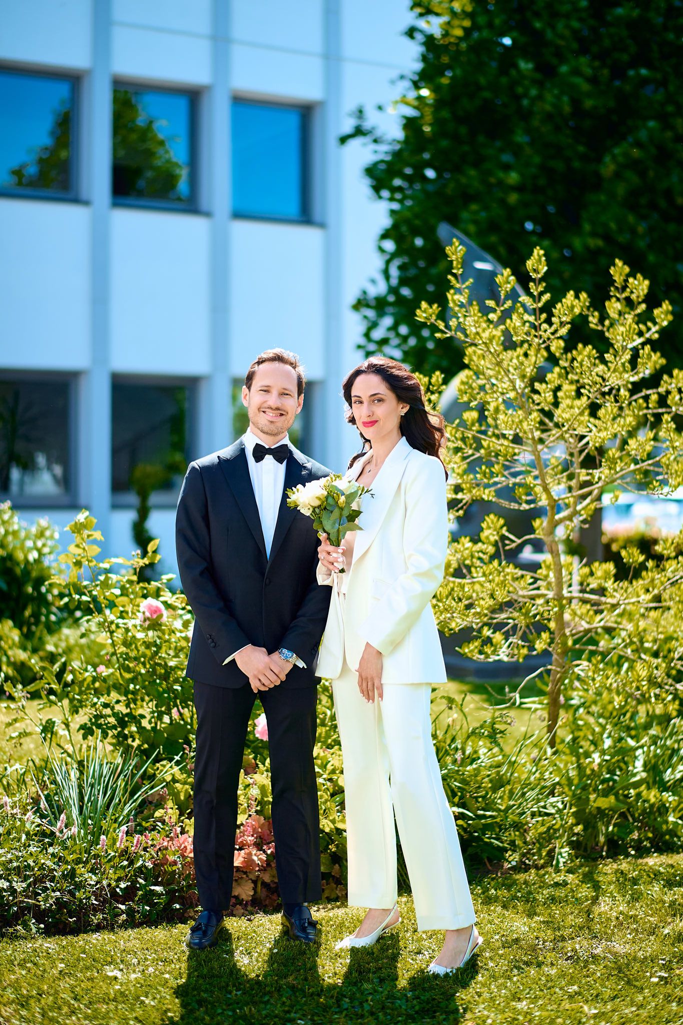 Couple in formal attire poses together in a sunny garden with yellow flowers.