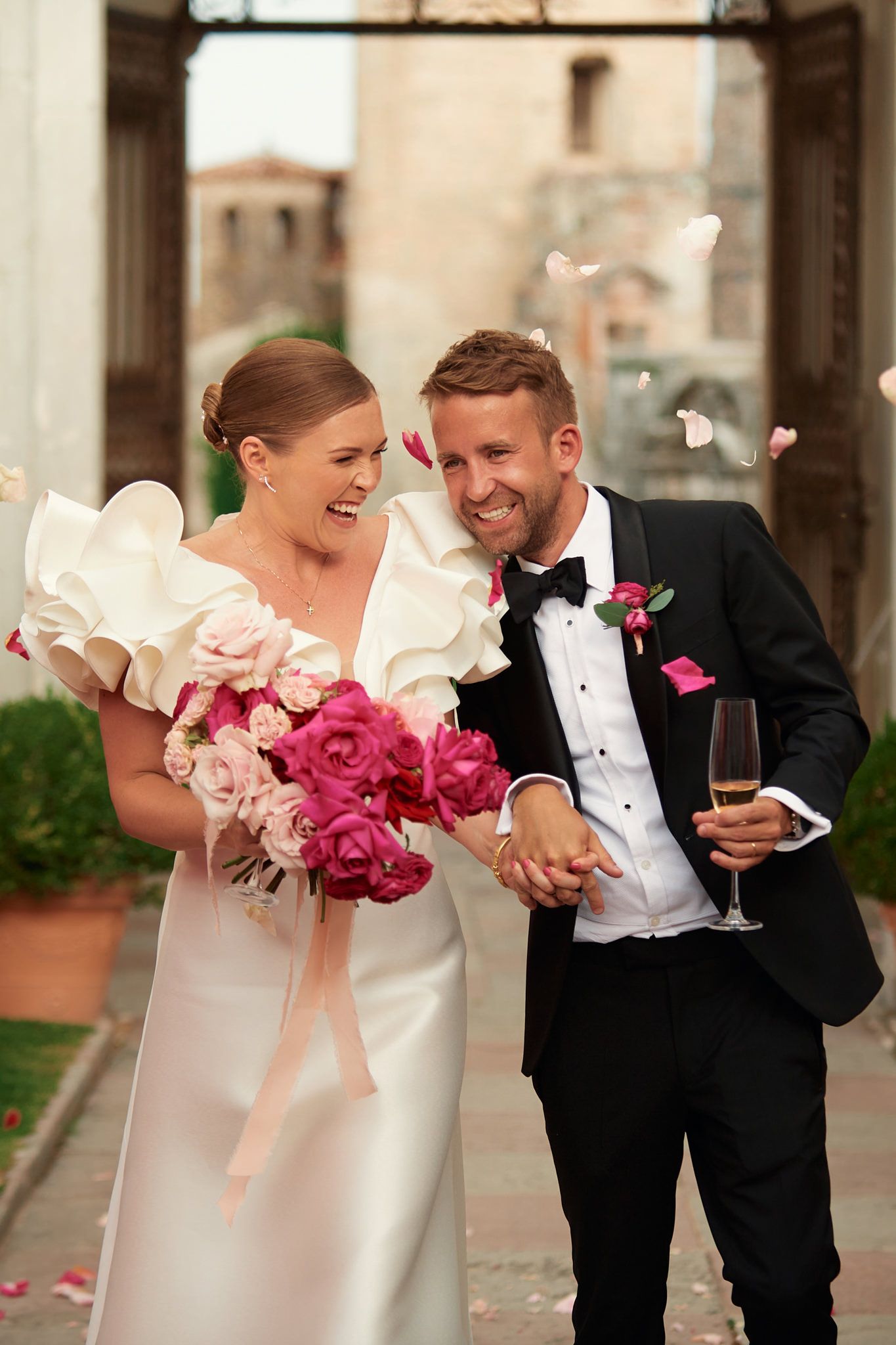 Couple in formal attire sharing laughter while holding vibrant pink bouquets.
