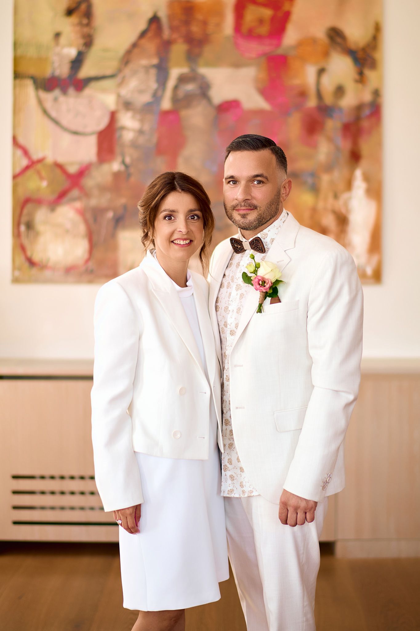 Wedding couple poses in matching white outfits against abstract art gallery backdrop.