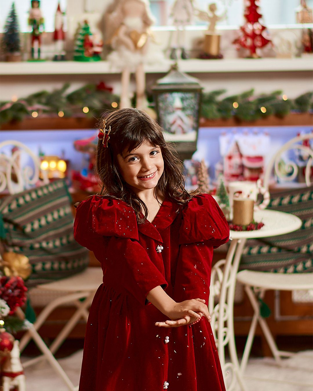 A smiling girl in a red velvet dress poses among festive Christmas decorations and holiday figurines.