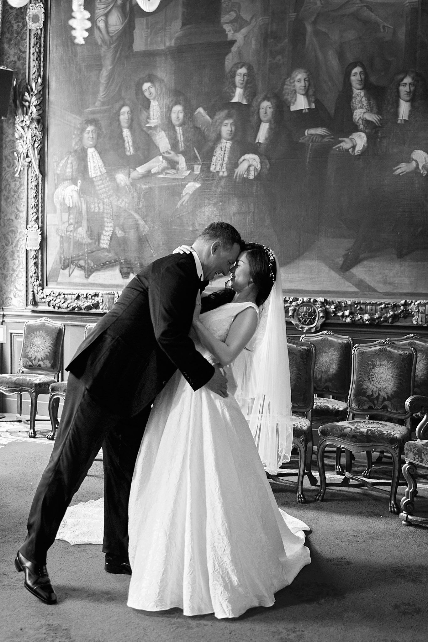 A romantic black and white wedding photo of a couple kissing in an elegant room with a historic painting backdrop.