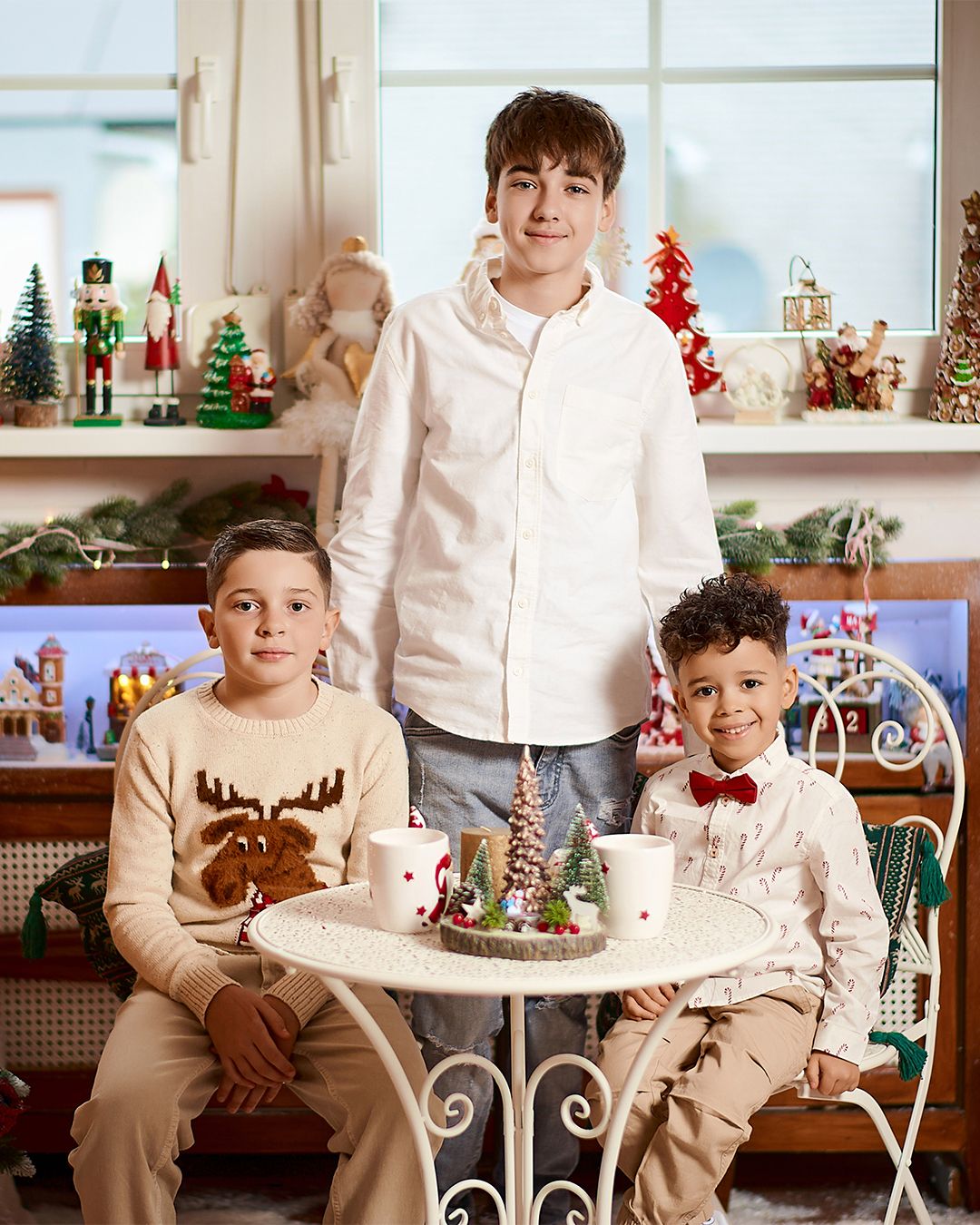 Three boys pose for a festive Christmas photo surrounded by holiday decorations, nutcrackers, and a small decorated table.