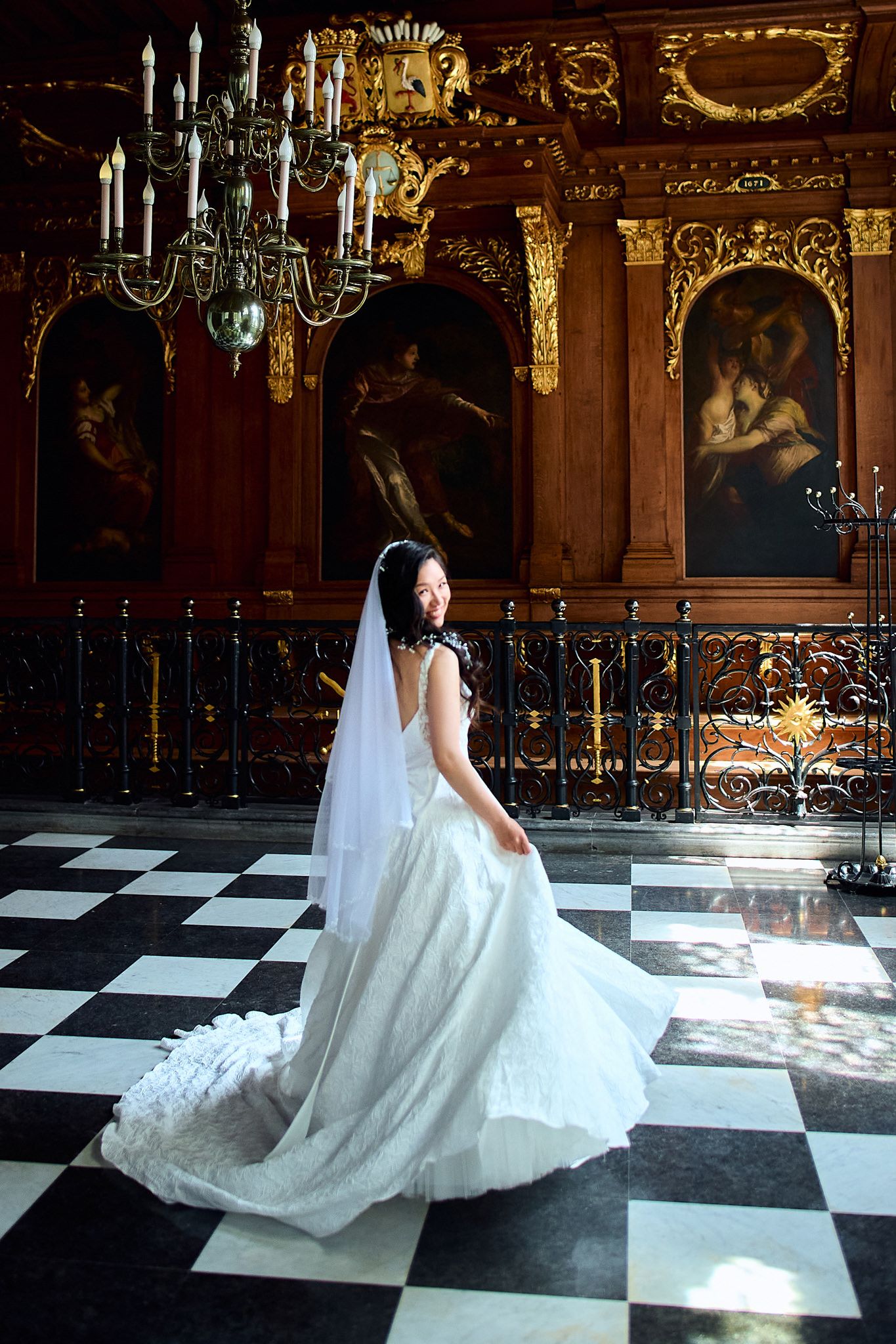 Bride in flowing white gown poses on black and white checkered floor under elegant chandelier.
