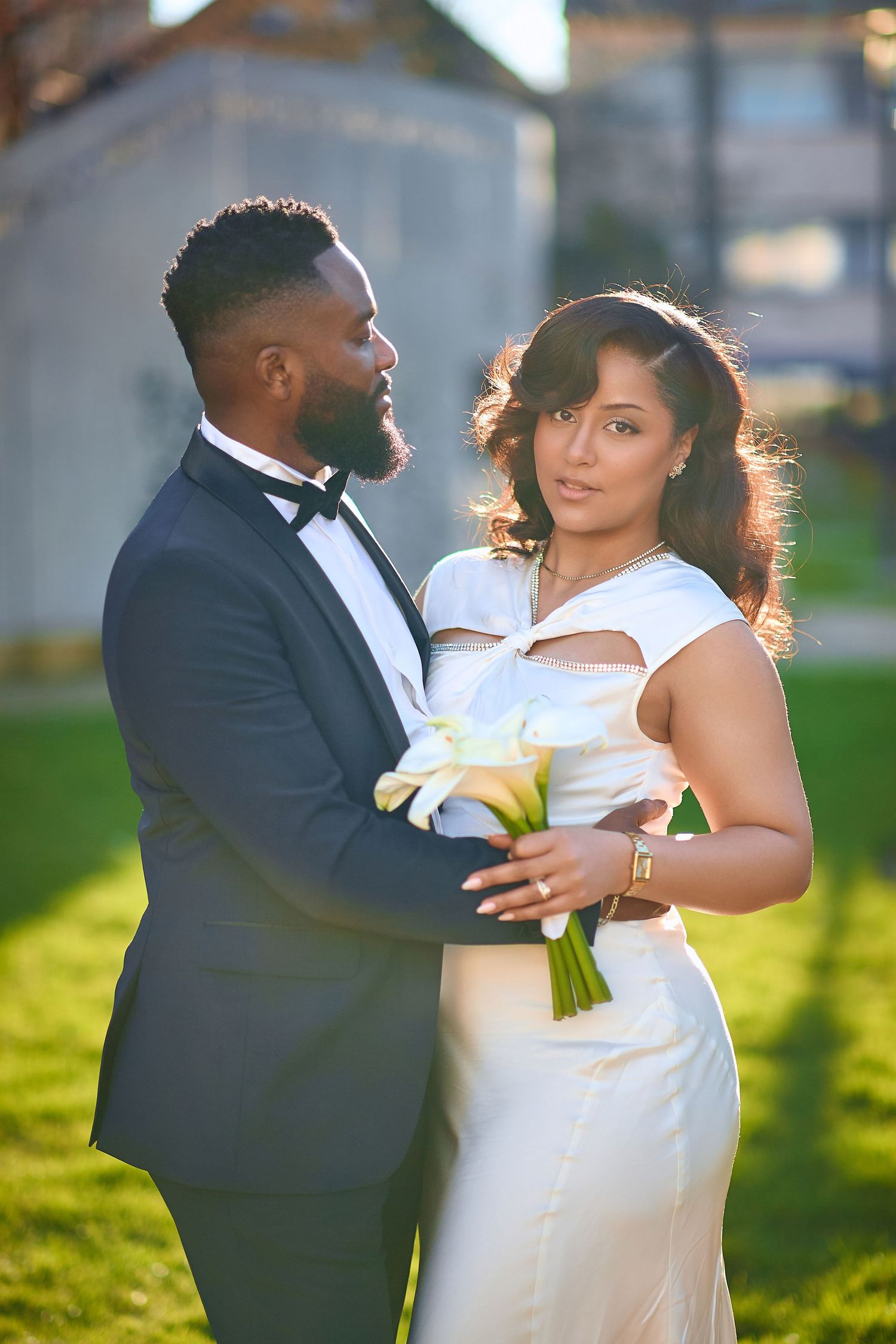 A couple in wedding attire dances together on a sunlit grassy lawn during their romantic outdoor photoshoot.