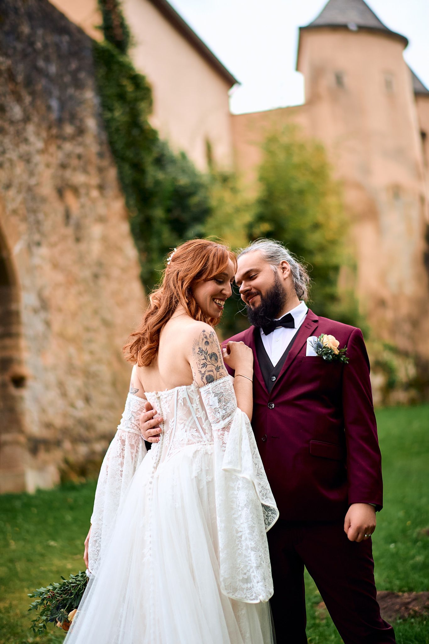 Wedding couple shares tender moment near castle walls in burgundy and white attire.