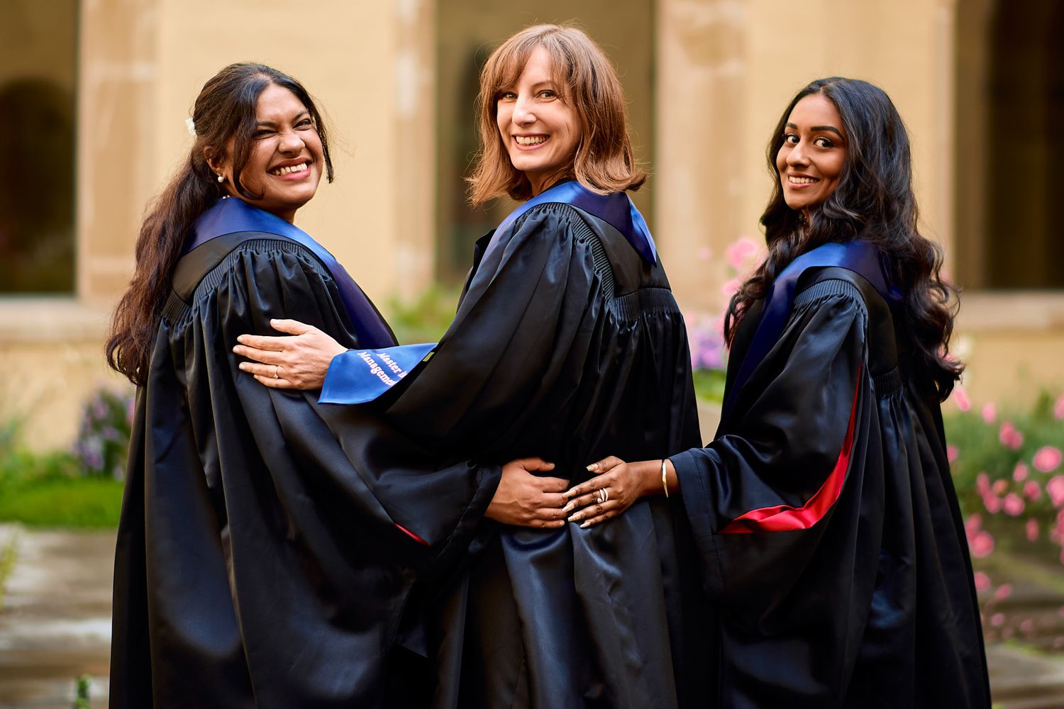 Graduates in traditional academic regalia hug and celebrate outdoors.
