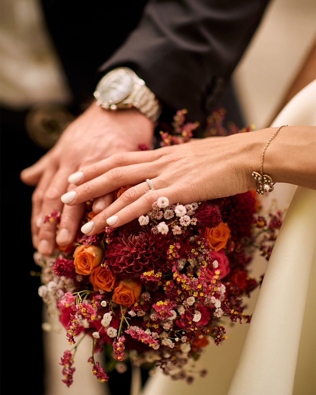 Newlyweds hands resting on a vibrant wedding bouquet with red, orange, and pink flowers, showcasing wedding rings.