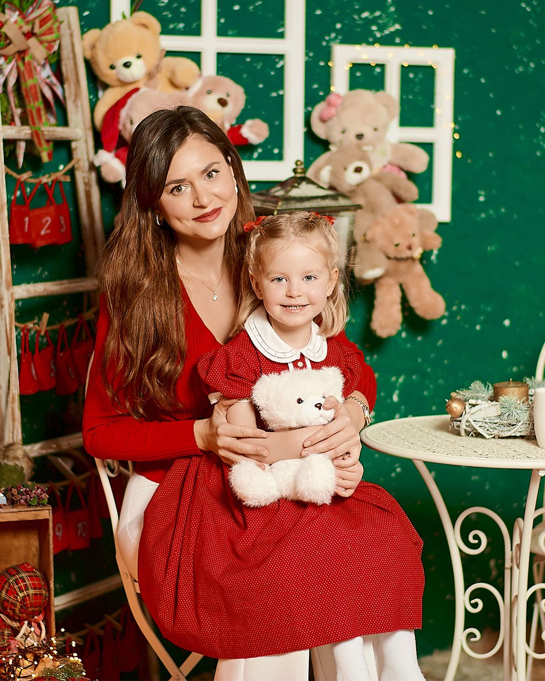 Woman in red dress holds smiling girl with white teddy bear in festive Christmas photo studio with green backdrop.