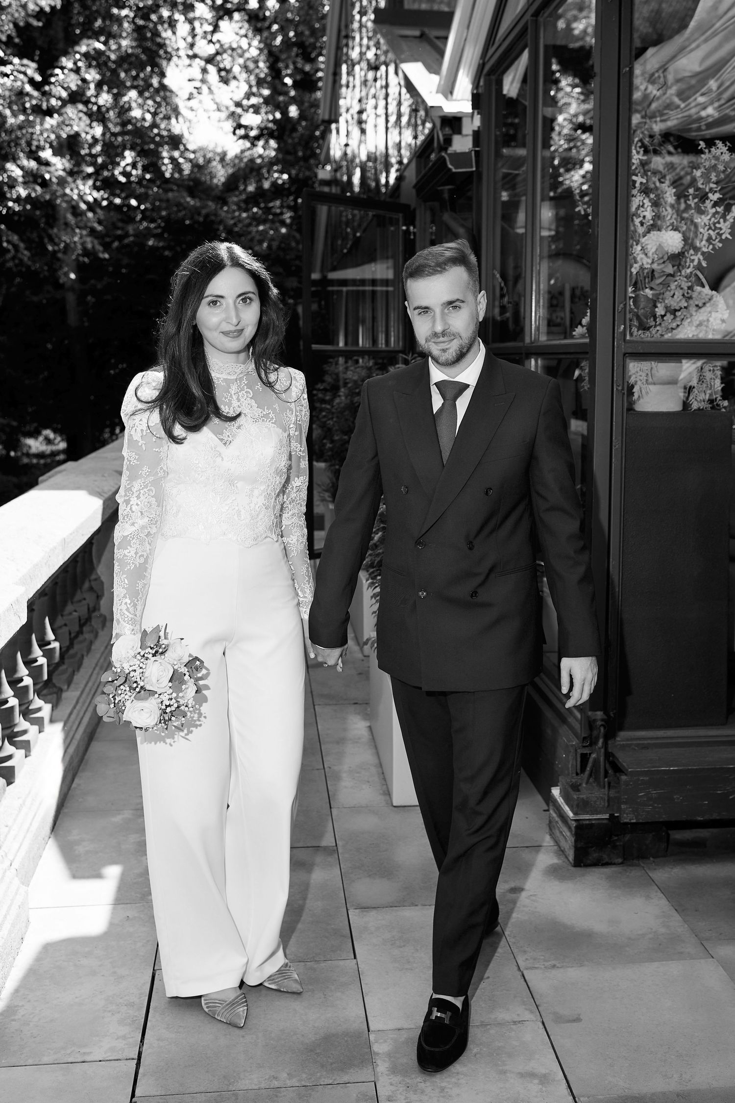 Black and white photo of a bride in white lace jumpsuit and groom in dark suit holding hands outdoors.
