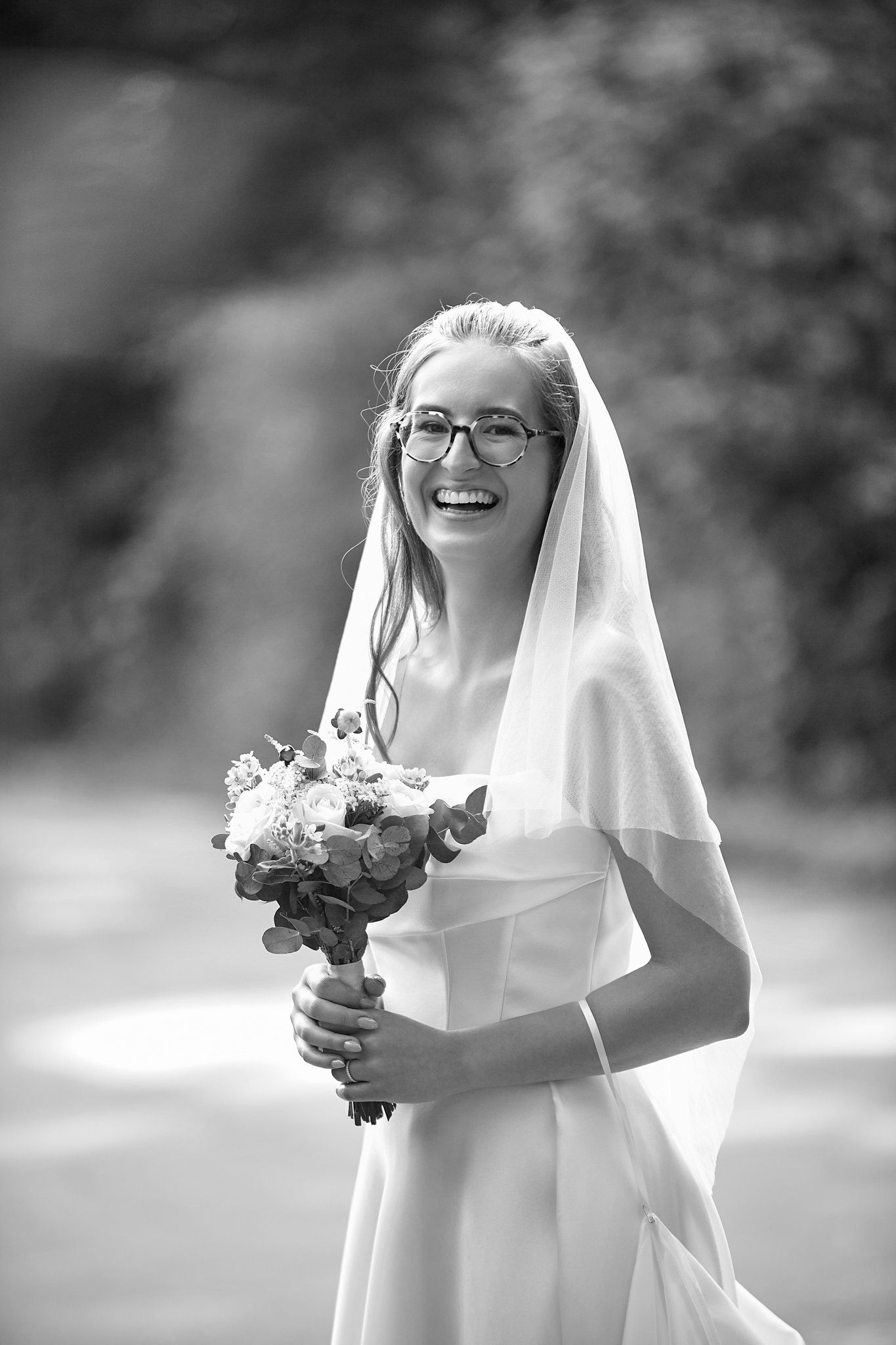 Black and white portrait of bride in glasses holding bouquet and laughing.