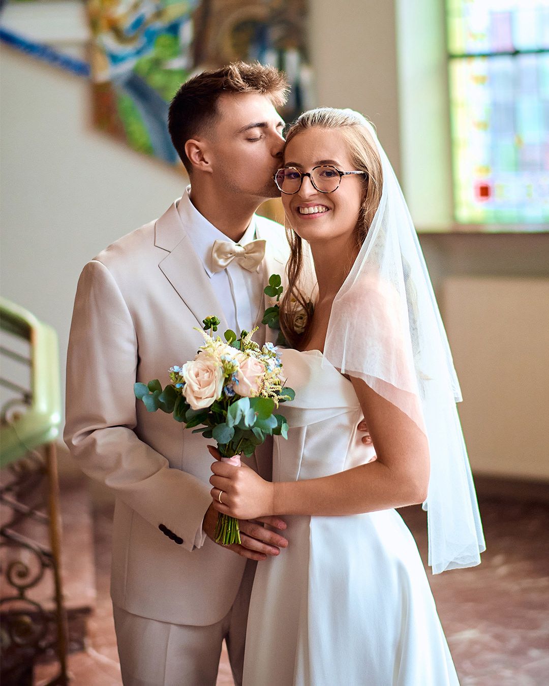 A wedding couple shares a sweet moment in a church with stained glass windows and pink flowers.
