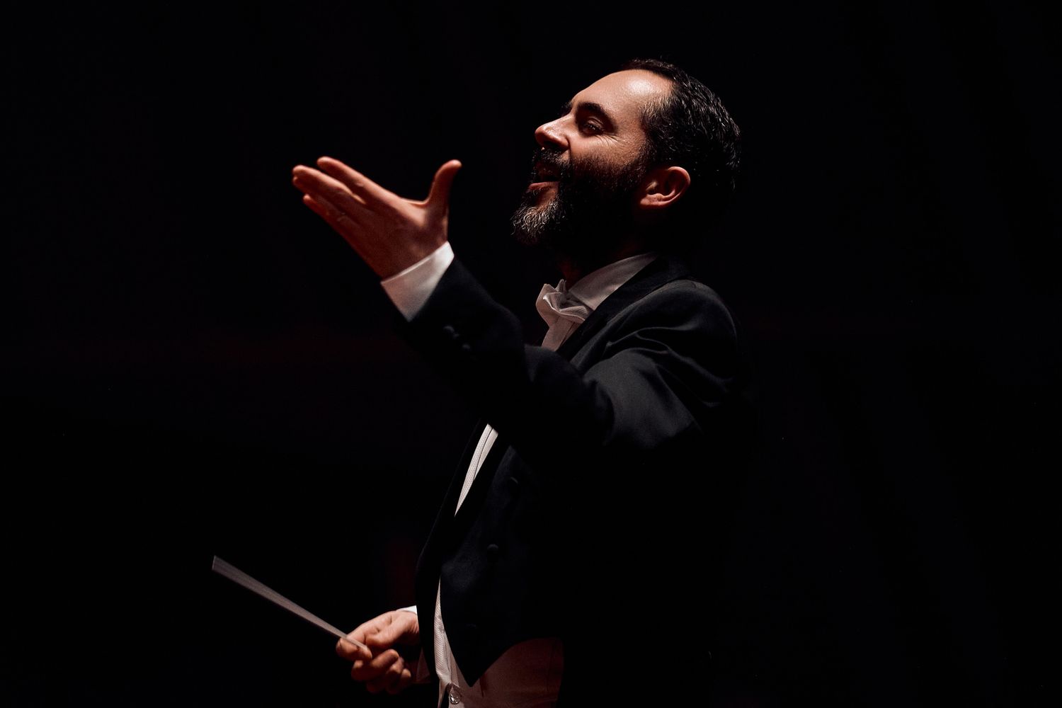 Orchestra conductor in tuxedo expressively directs with baton against black background.