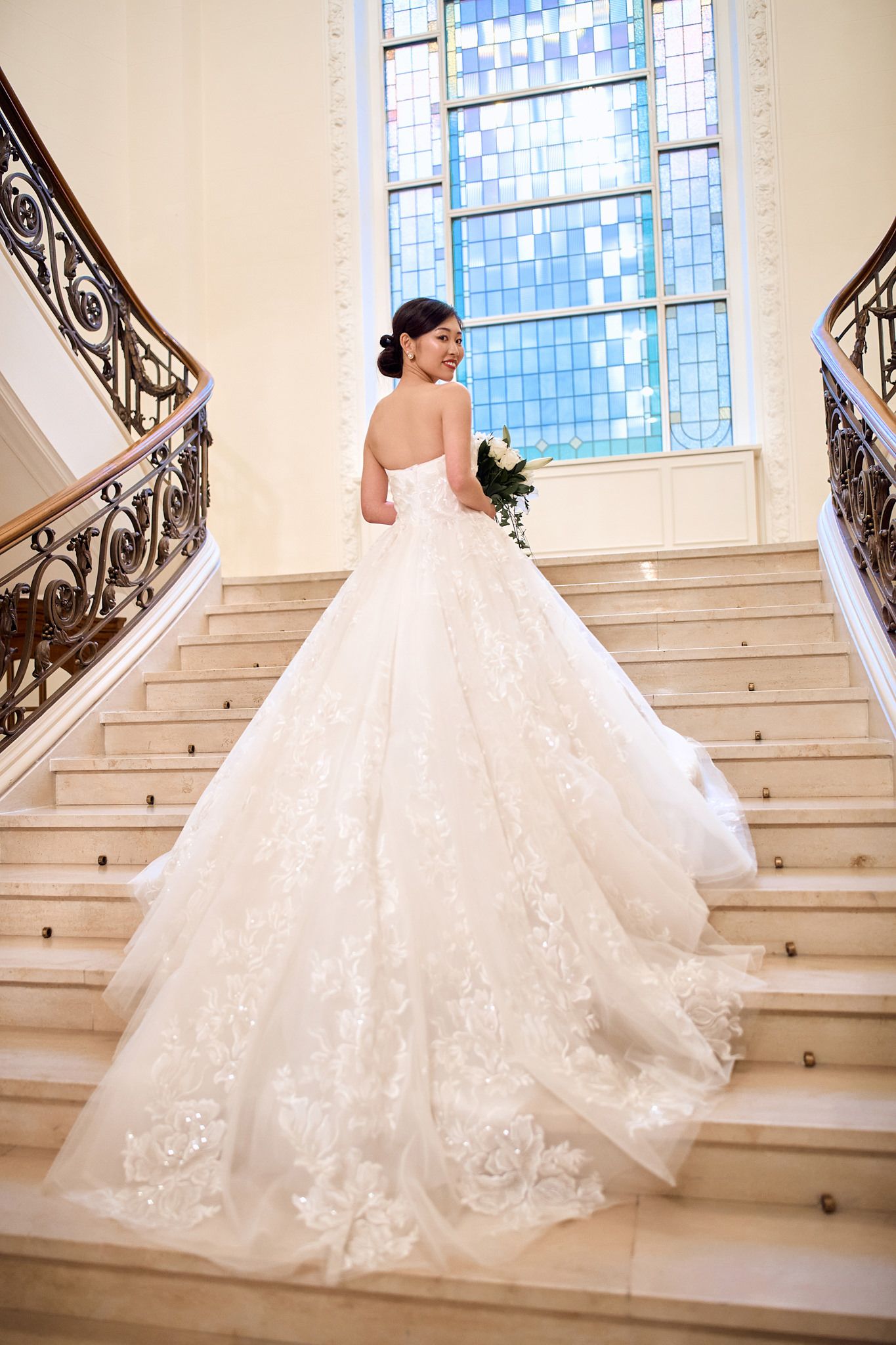 A bride in an elegant white lace gown descends a grand staircase with ornate railings and stained glass windows.