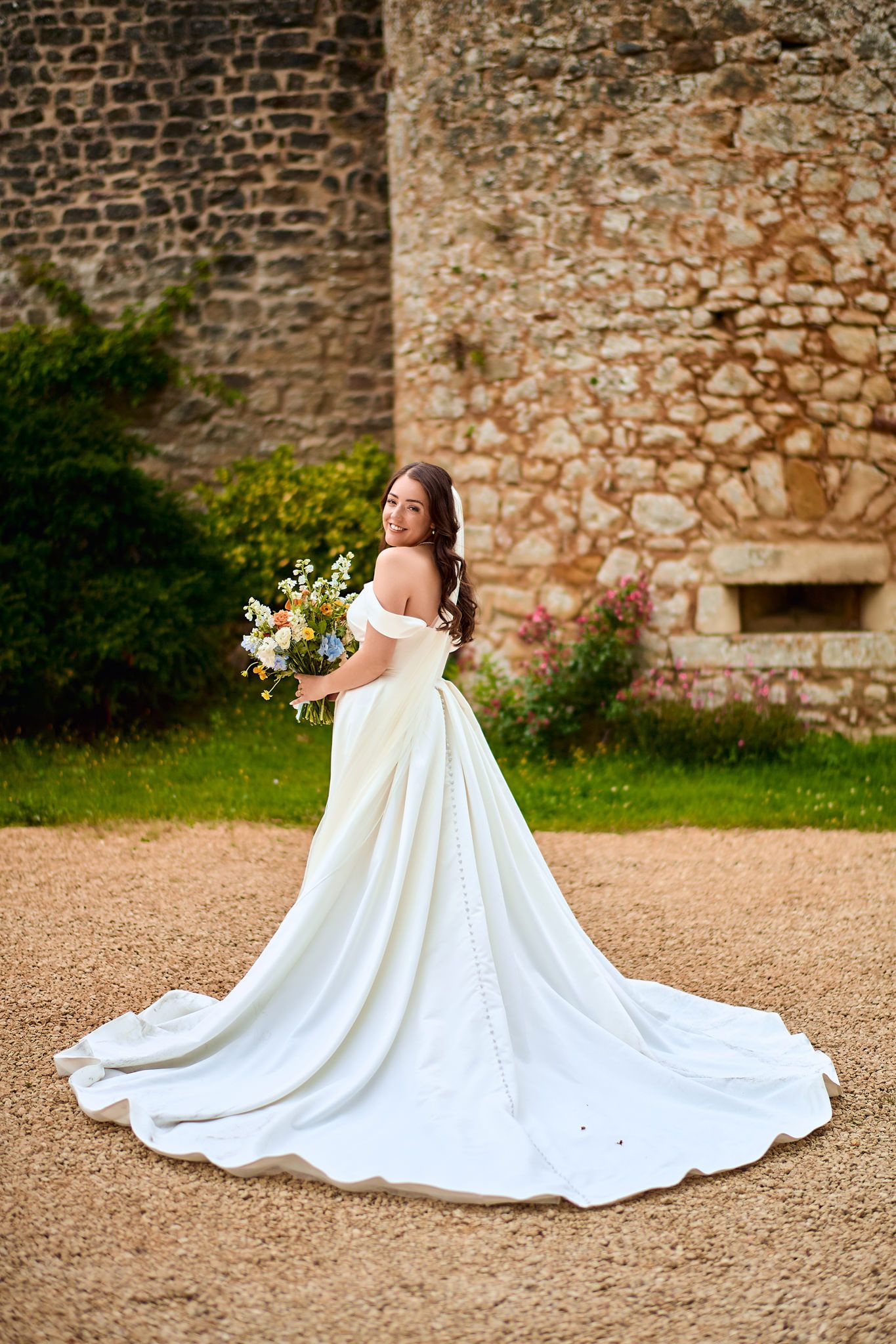 A flowing white wedding dress with a dramatic train spread out against a stone wall backdrop.