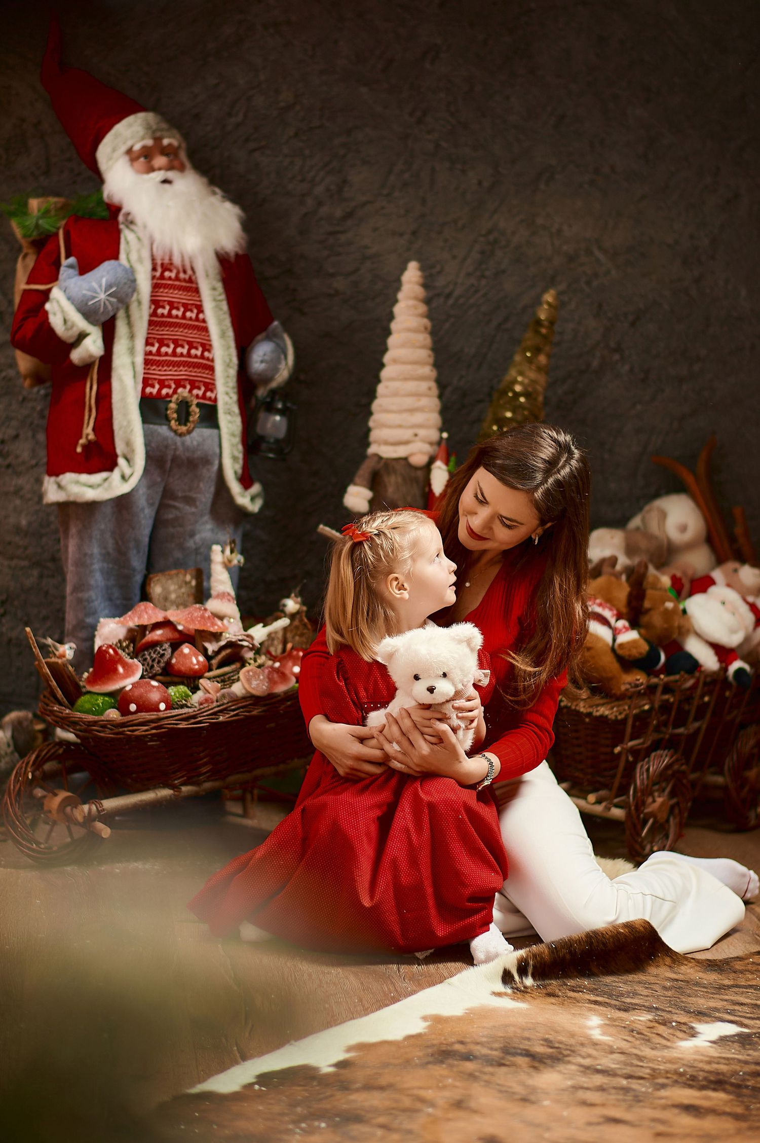 A mother and young daughter in red dresses share a tender moment with a white teddy bear in a festive Christmas setting.