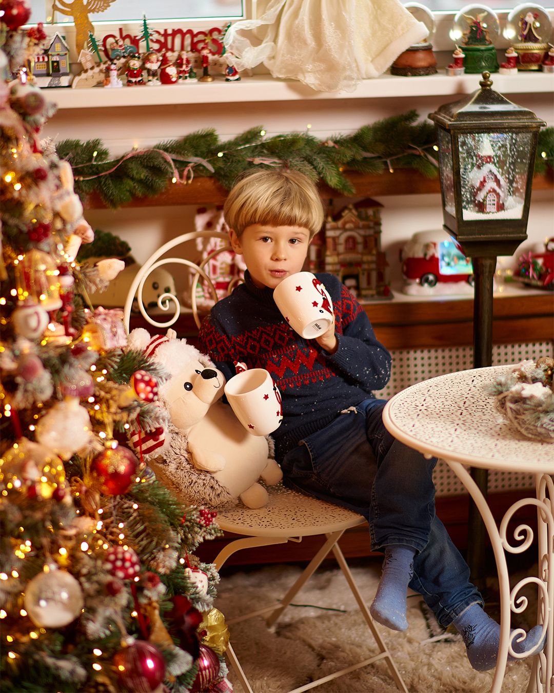 Young boy sitting by a decorated Christmas tree holding festive toys in a holiday-themed room with lights and ornaments.