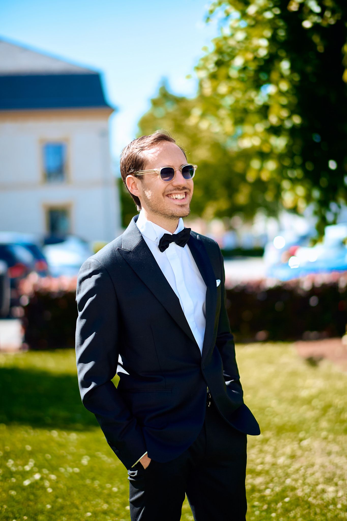 A person in a black tuxedo and sunglasses poses outdoors on a sunny day with buildings in the background.
