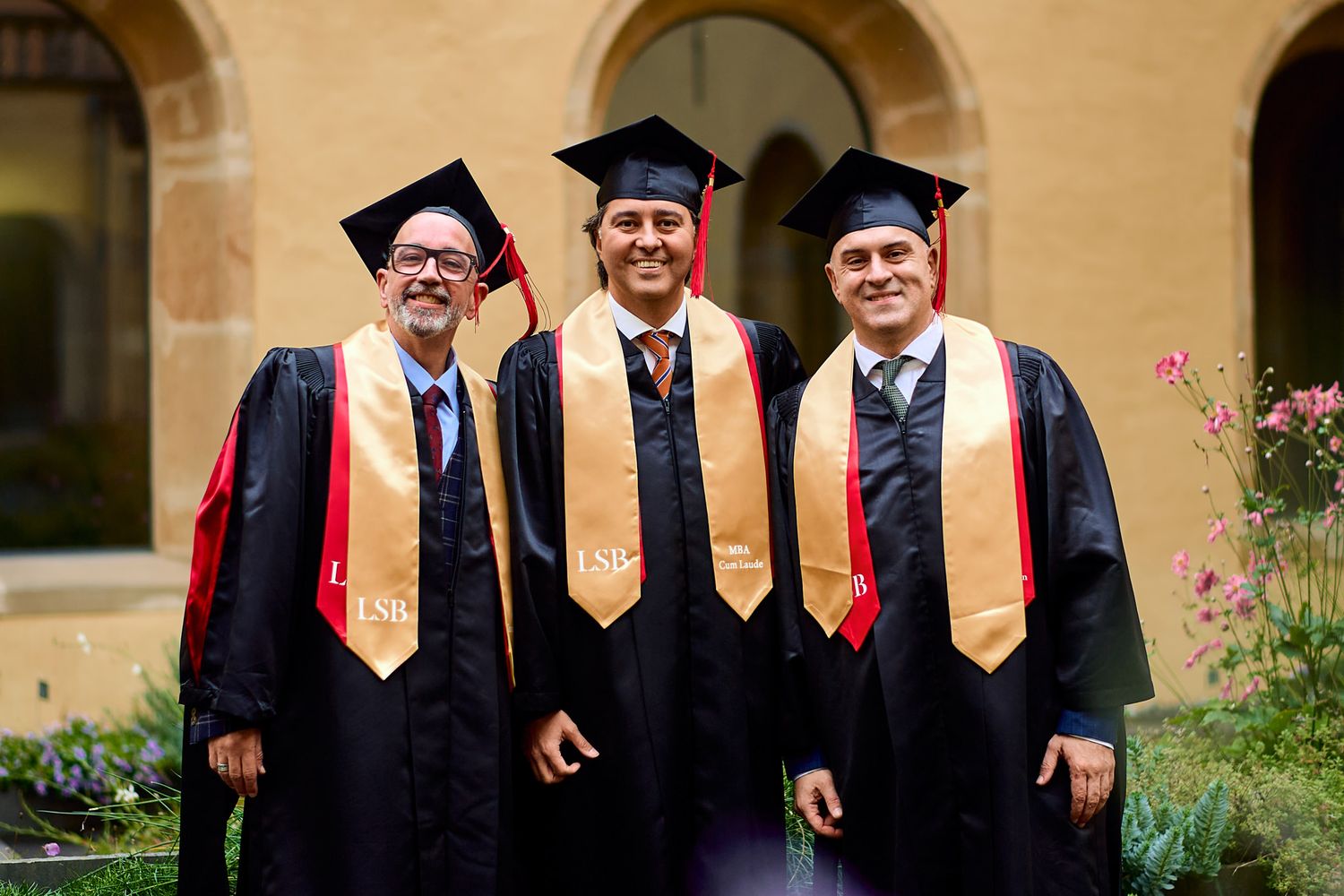 Three graduates wearing black robes and orange stoles pose together at commencement ceremony.