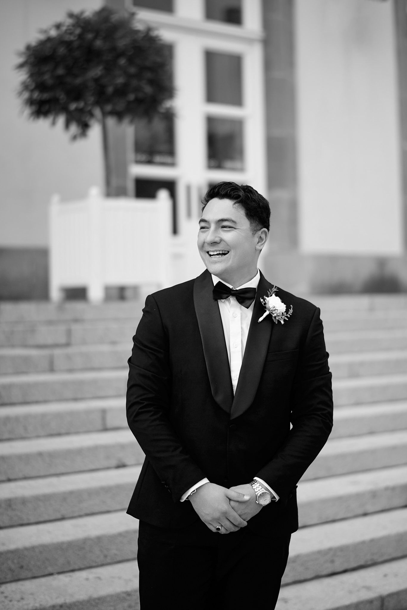 A groom in a black tuxedo smiles and laughs while standing on stone steps outside a building.