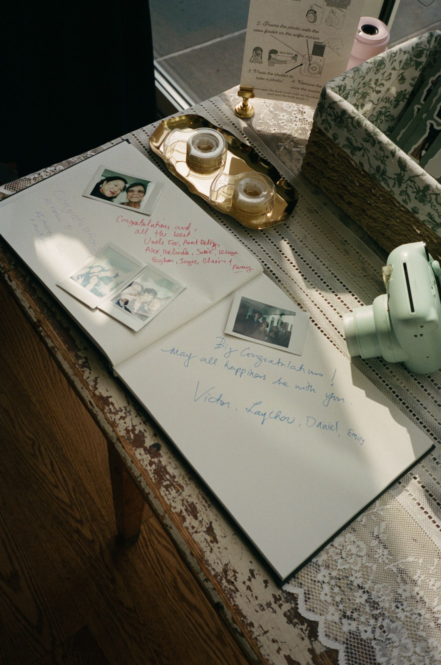 A rustic table displays a guest book with handwritten notes, Polaroid photos, tape, and decorative items in warm light.