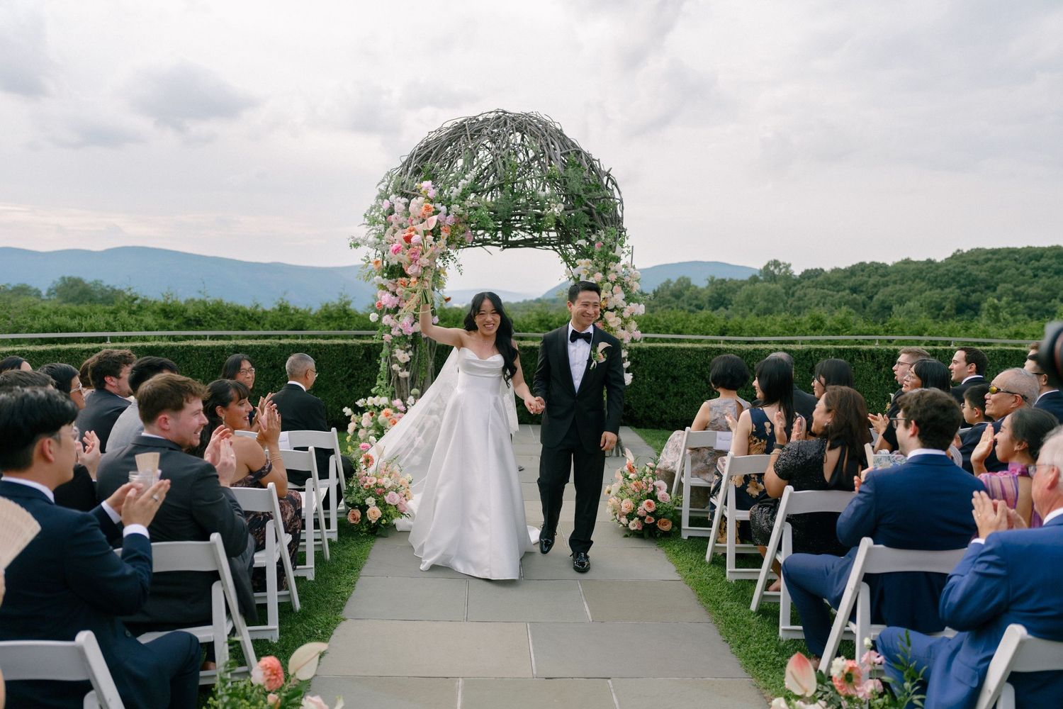 Bride and groom walk down outdoor aisle flanked by guests, floral arch, and scenic mountain views.