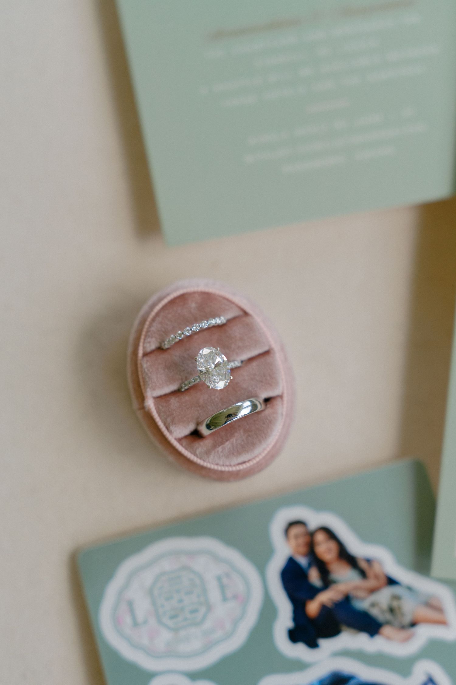 Oval pink velvet ring box holding diamond engagement ring, wedding band, and eternity band beside wedding stationery.