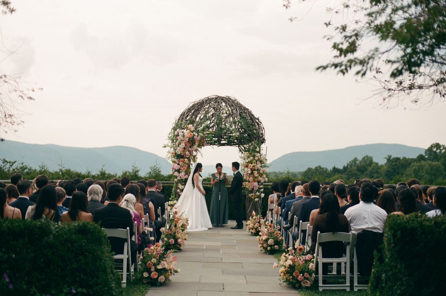 Outdoor wedding ceremony with floral arch, mountain backdrop, guests seated on white chairs along a petal-lined aisle.