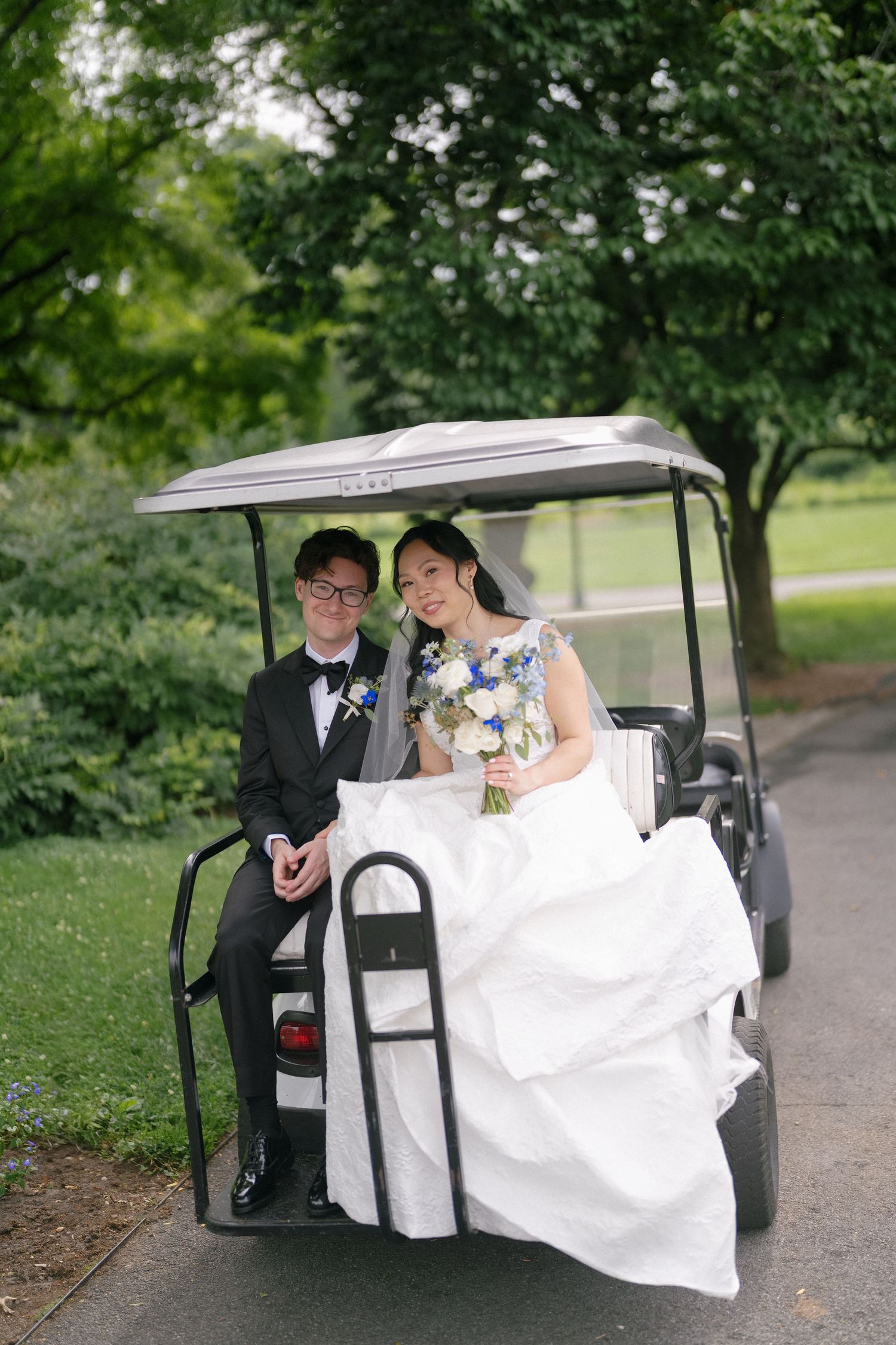 Smiling bride and groom ride a golf cart through a lush green botanical garden on their wedding day.
