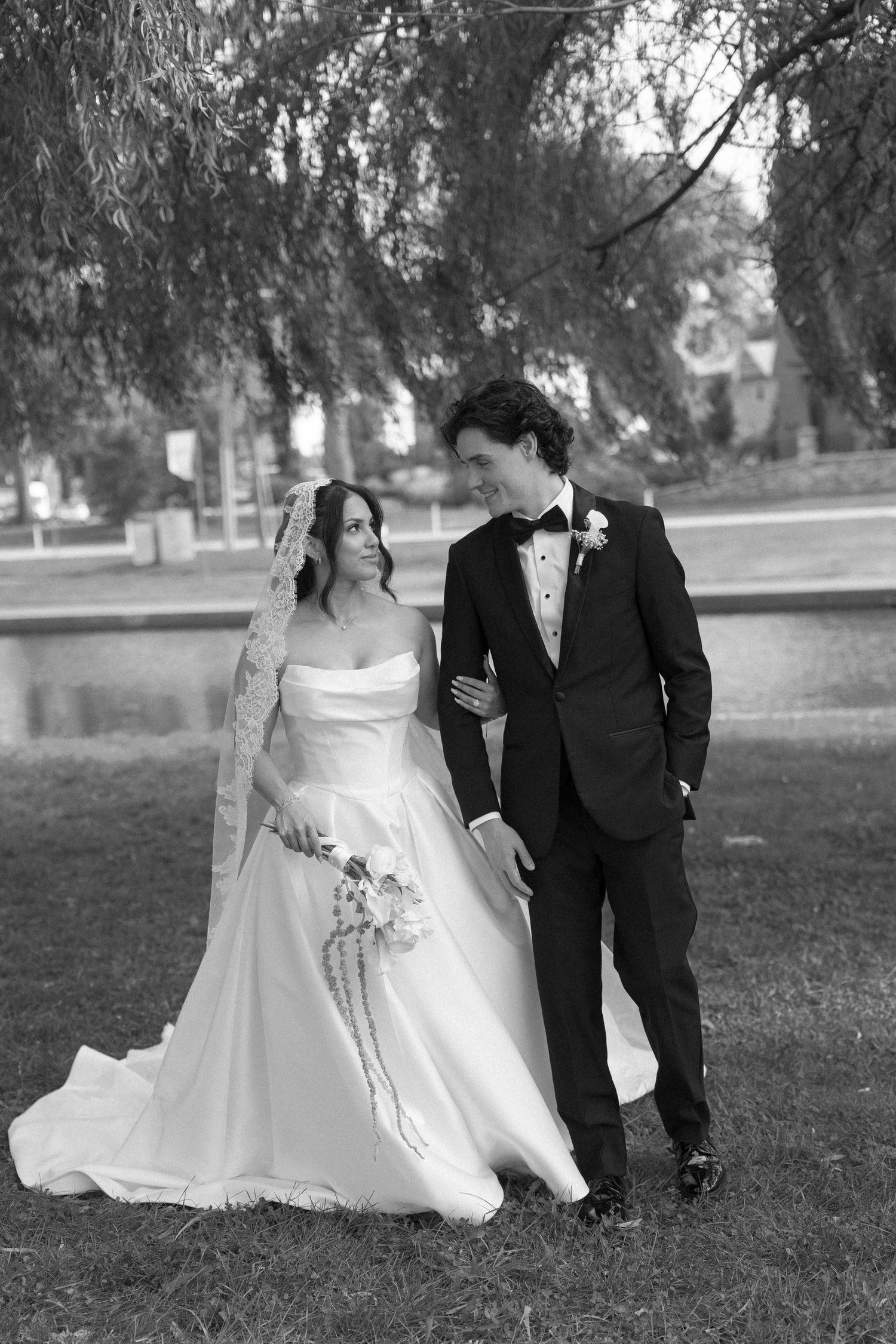 Black and white photo of bride and groom walking together under willow tree by a pond on wedding day.